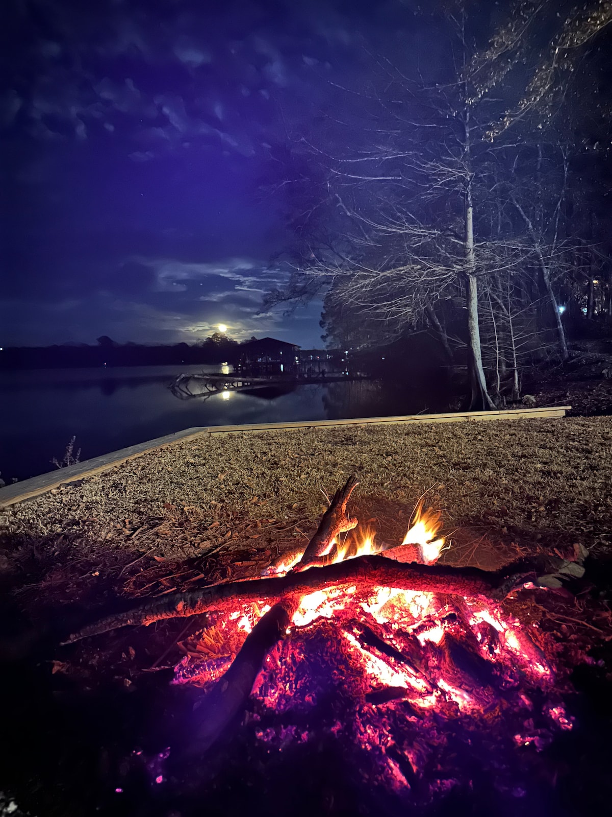 A campfire glows warmly at the lakeside, with orange and red flames rising over darkened logs. The tranquil lake reflects a twilight sky, while silhouettes of nearby trees and a distant cabin can be seen, creating a serene nighttime environment.