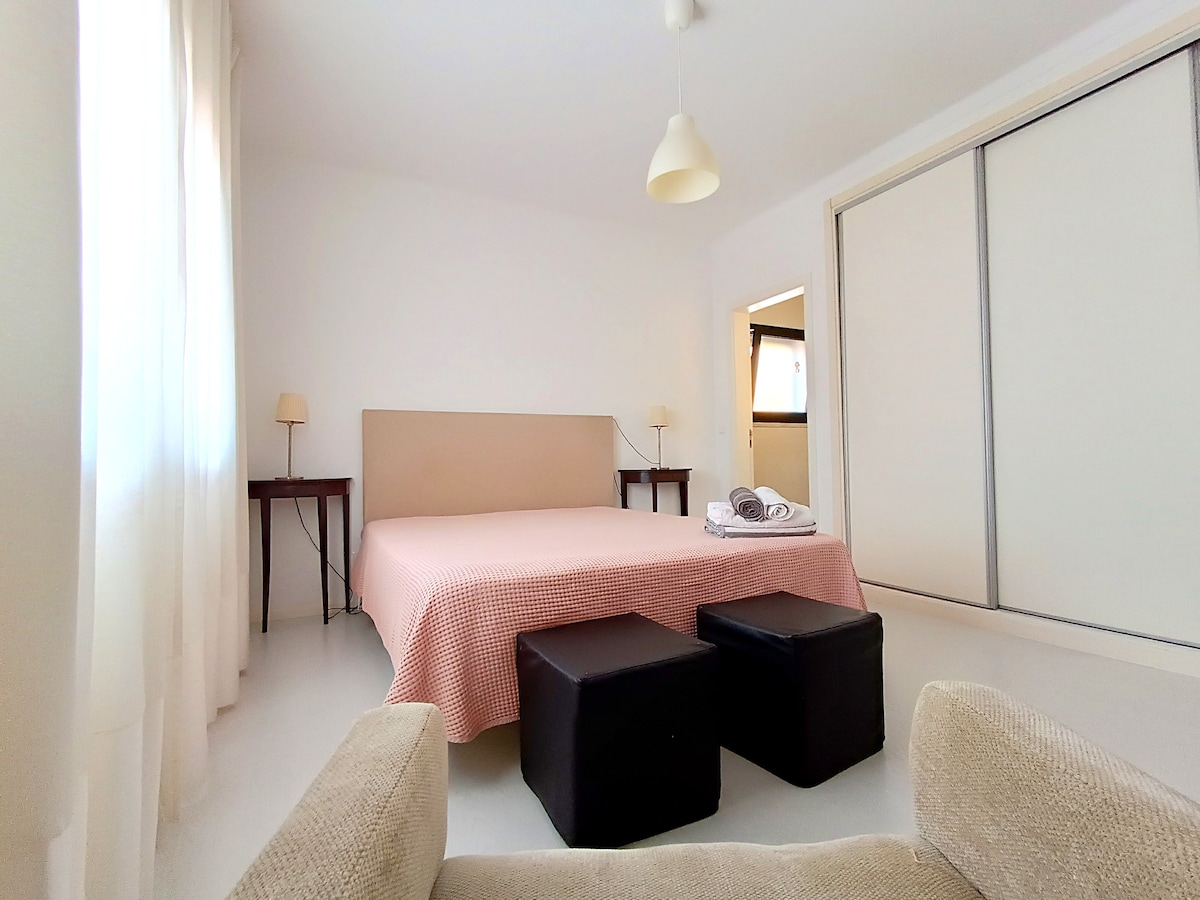 A spacious room is presented with a light-colored wall and a large bed covered in a soft quilt. Two nightstands flank the bed, while a comfortable sofa and two ottomans provide additional seating. Natural light fills the space through sheer curtains.