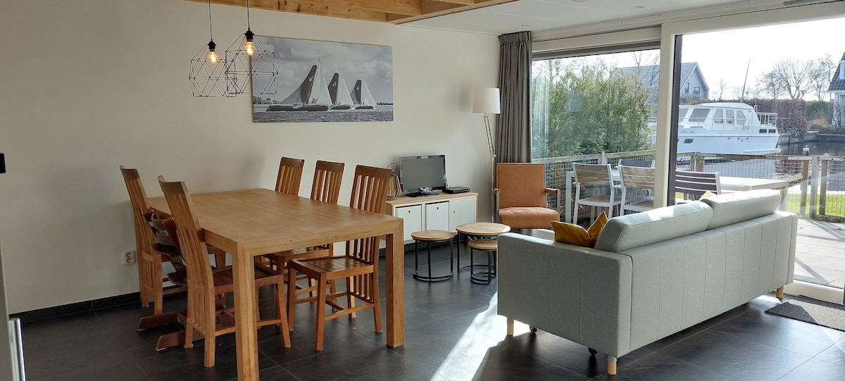 A spacious living area is featured, equipped with a large wooden dining table and six chairs. A comfortable sofa is positioned nearby, complemented by a side table. Natural light floods the space through expansive windows, revealing views of the outdoor area.