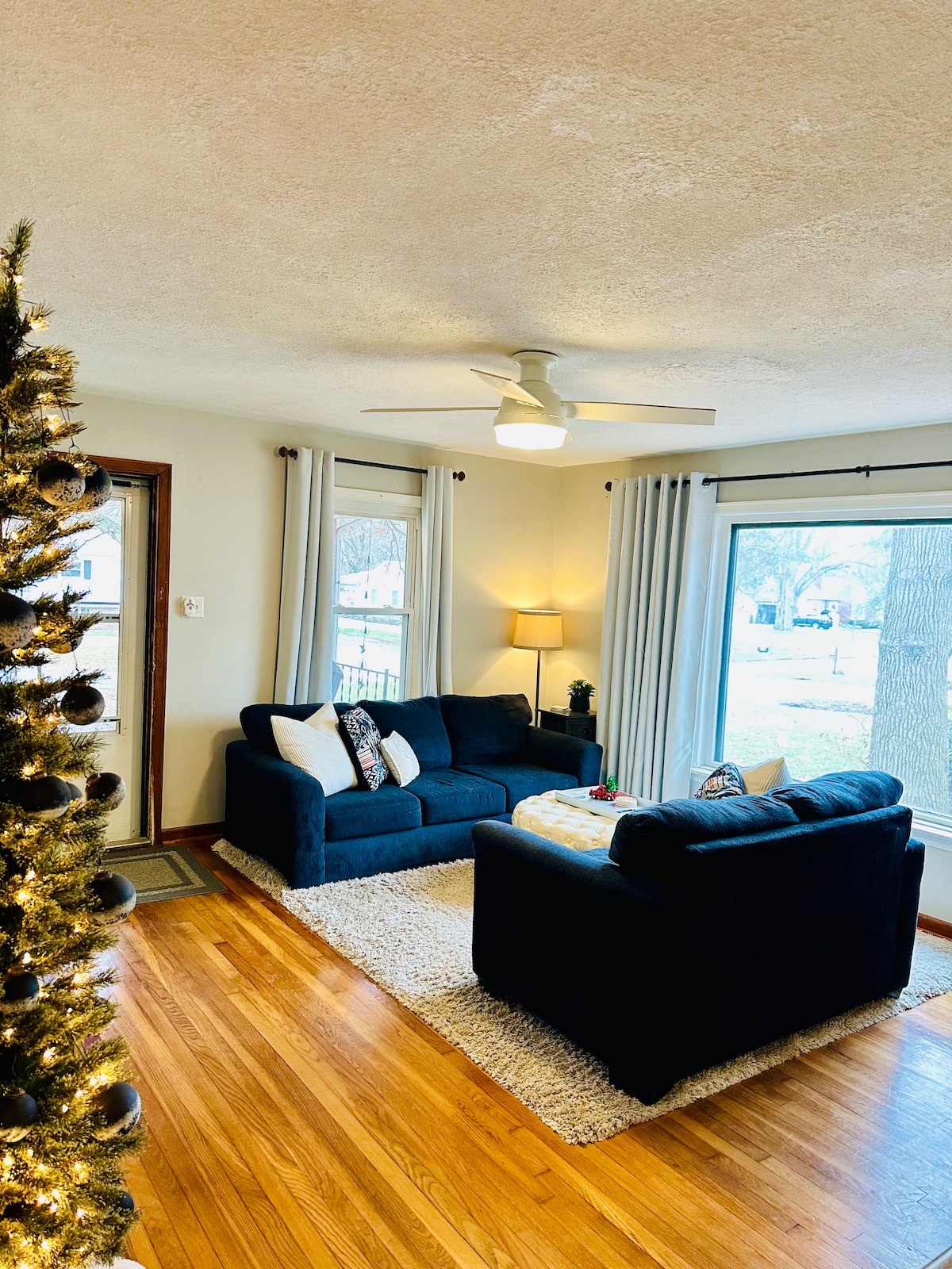 A living room showcases two dark sofas positioned around a low table, with natural light streaming through large windows. A cozy area rug lies beneath, and a festive tree is visible in the corner, adding a touch of warmth to the space.