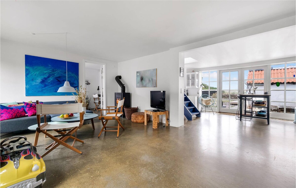 An open living space is highlighted by a concrete floor and large windows allowing natural light. A fireplace is positioned near the seating area, which features colorful cushions. A dining table is surrounded by chairs, providing a casual setting in this airy environment.