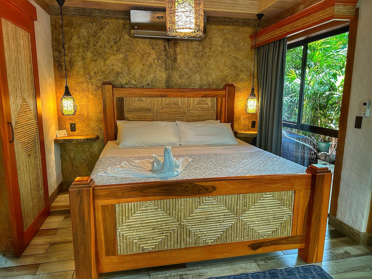 A spacious king bed with a textured headboard is centered in the well-lit room. Soft lighting is provided by pendant lamps hanging from the ceiling, while large windows reveal a view of lush greenery. Natural materials are showcased in the wooden furnishings.