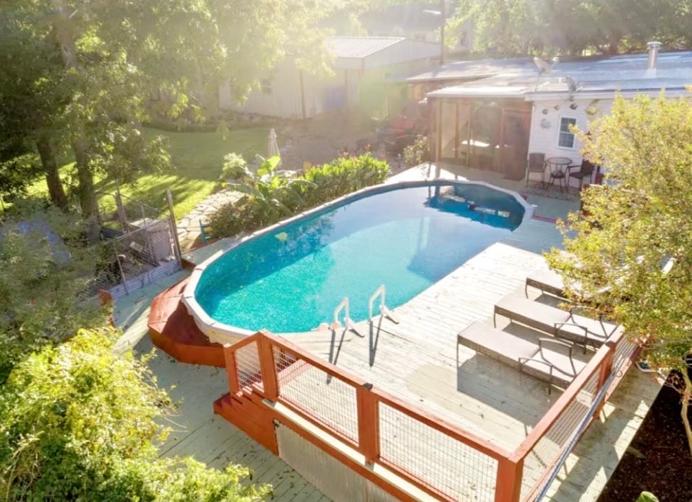 An above-ground pool is set in a serene outdoor space, surrounded by lush greenery. A wooden deck offers lounge chairs for relaxation. A screened-in porch is visible in the background, enhancing the idyllic atmosphere. Sunlight reflects off the water, creating a peaceful setting.
