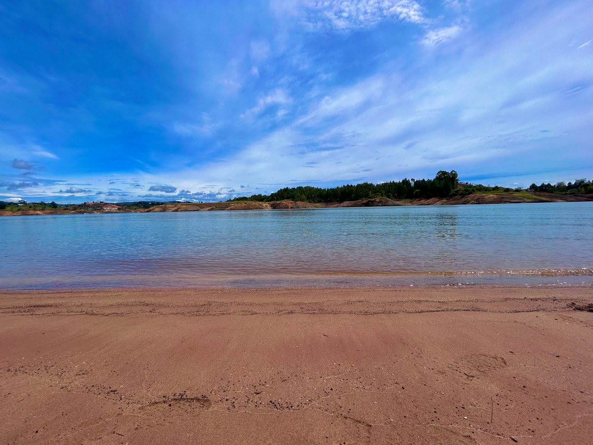 A serene view of a lake with calm waters reflecting the blue sky. The sandy shoreline stretches across the foreground, leading to gentle slopes of greenery in the distance. The scene conveys a sense of tranquility and connection to nature.