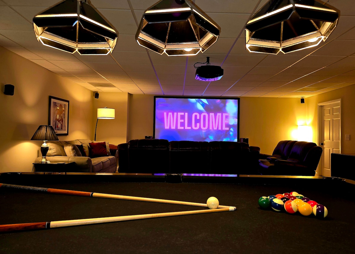 A spacious entertainment area features a pool table in the foreground, with colorful balls arranged on the felt. In the background, a large screen displays a welcome message, surrounded by comfortable seating and soft lighting from modern fixtures.