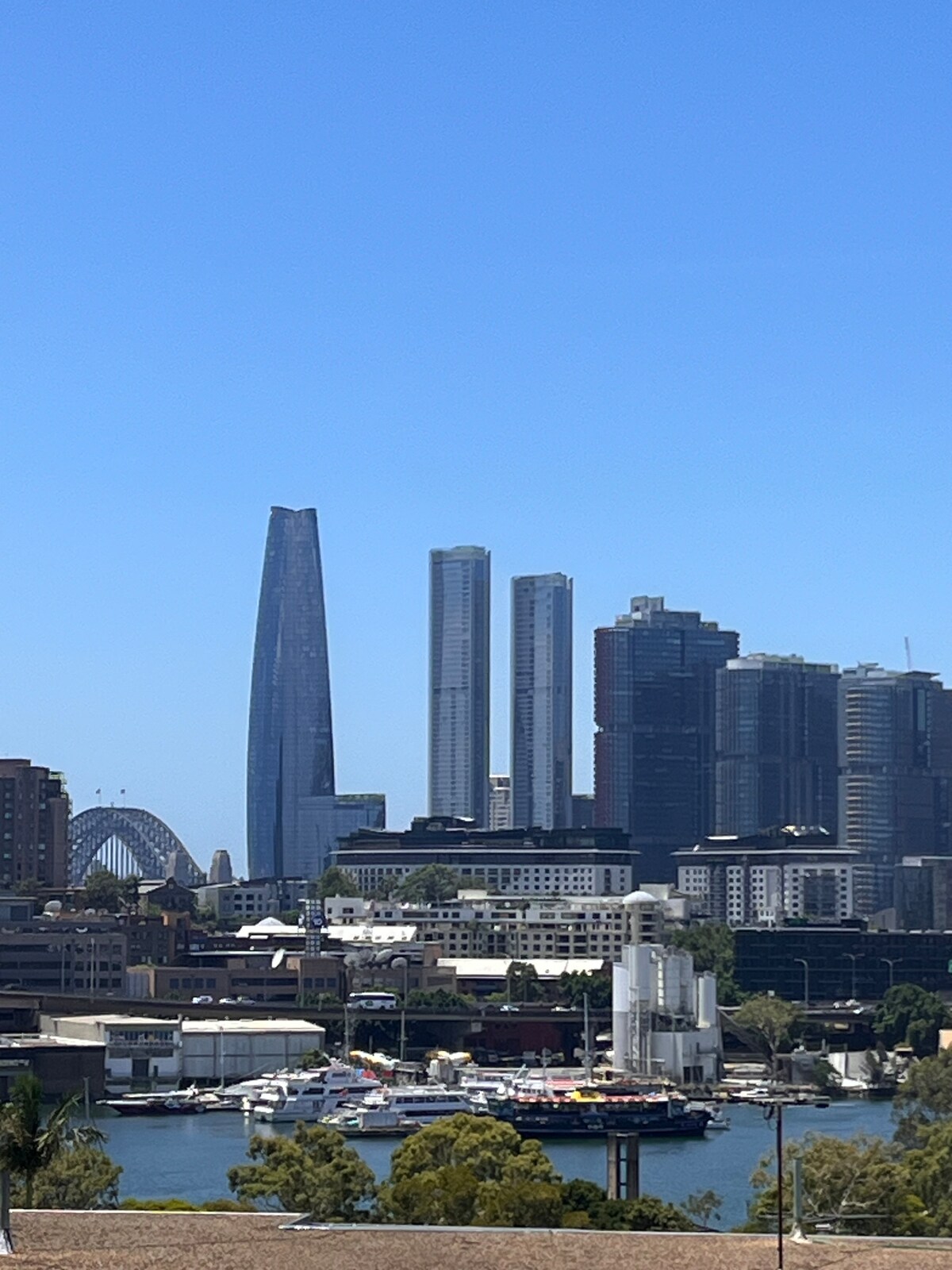 A clear daytime view of the Sydney skyline features modern skyscrapers, including a prominent tower and two additional tall buildings. The iconic Sydney Harbour Bridge is visible, alongside boats docked in the harbor, with greenery framing the foreground.