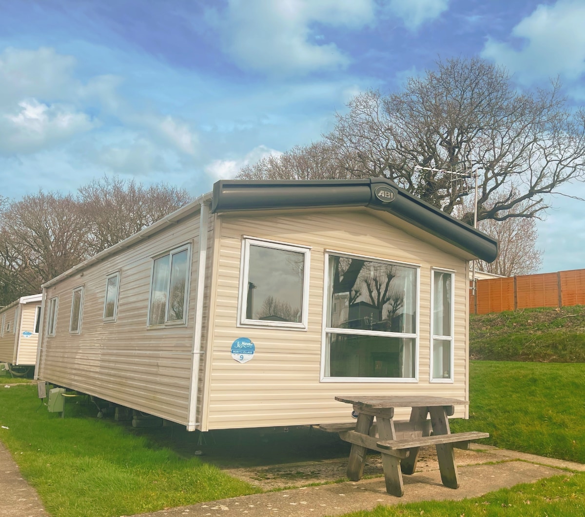 The exterior of a spacious caravan features large windows that provide ample natural light. Outdoor picnic seating is visible, positioned on a grassy area. Surrounding trees can be seen in the background under a partly cloudy sky.