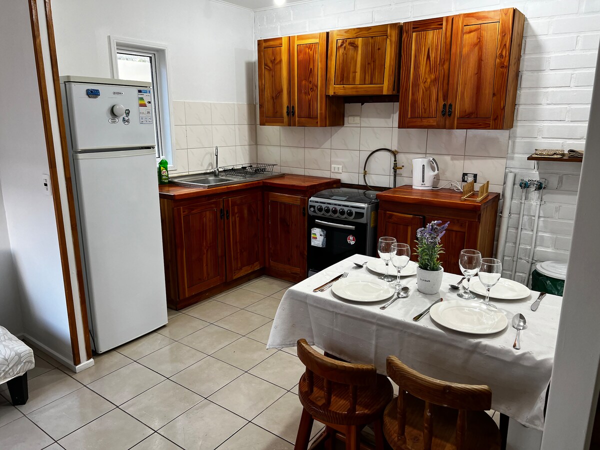 A functional kitchen features wooden cabinetry and a white tile backsplash. Appliances include a refrigerator, stove, and kettle. A dining table is set for four, adorned with glassware and a small potted plant, while the tile floor reflects the natural light from the window.