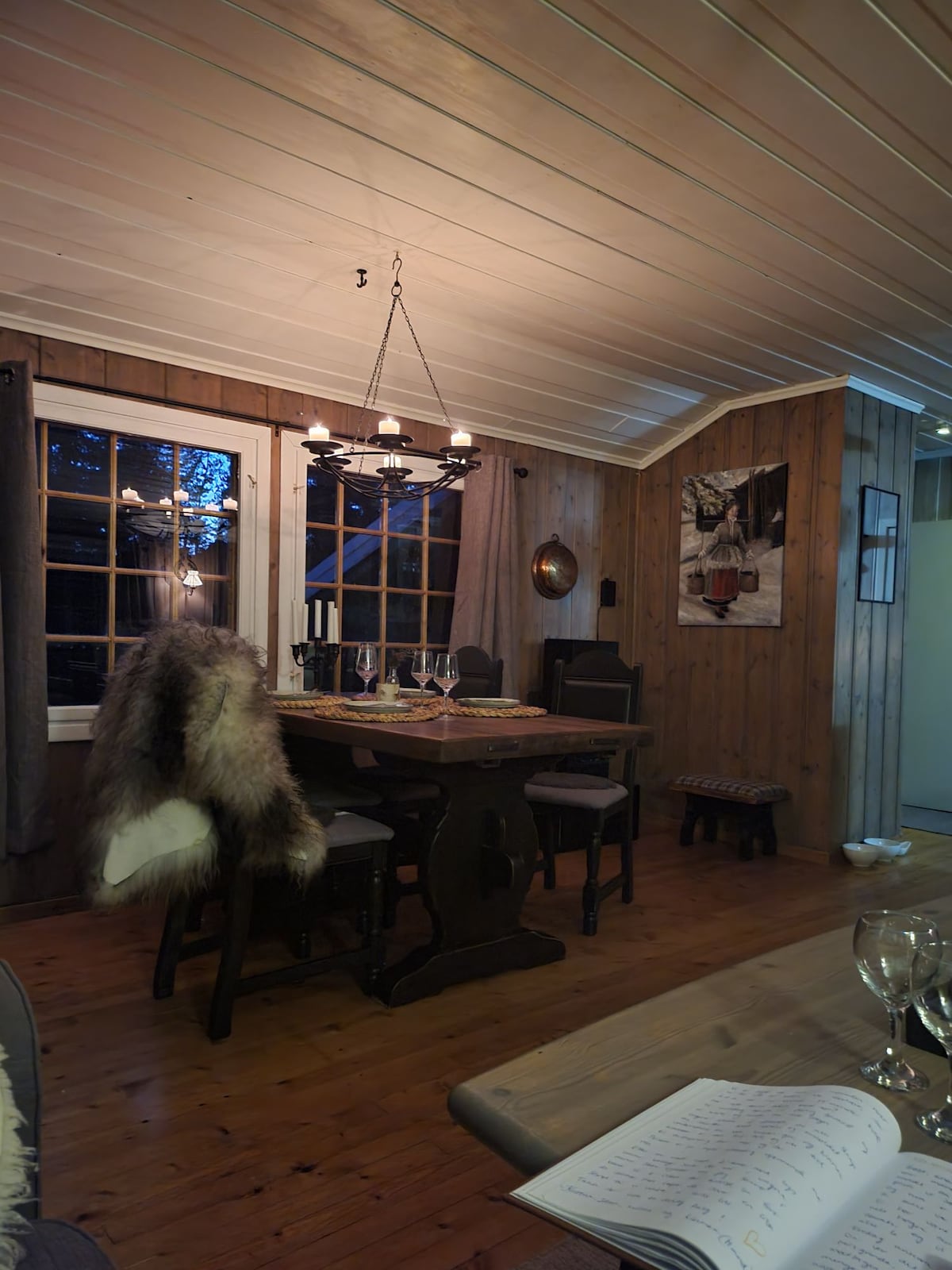 A dining area features a wooden table surrounded by chairs, illuminated by a chandelier. Large windows allow soft natural light to filter in, with a view of the exterior. A textured fur throw drapes over one chair, adding a cozy touch to the rustic space.