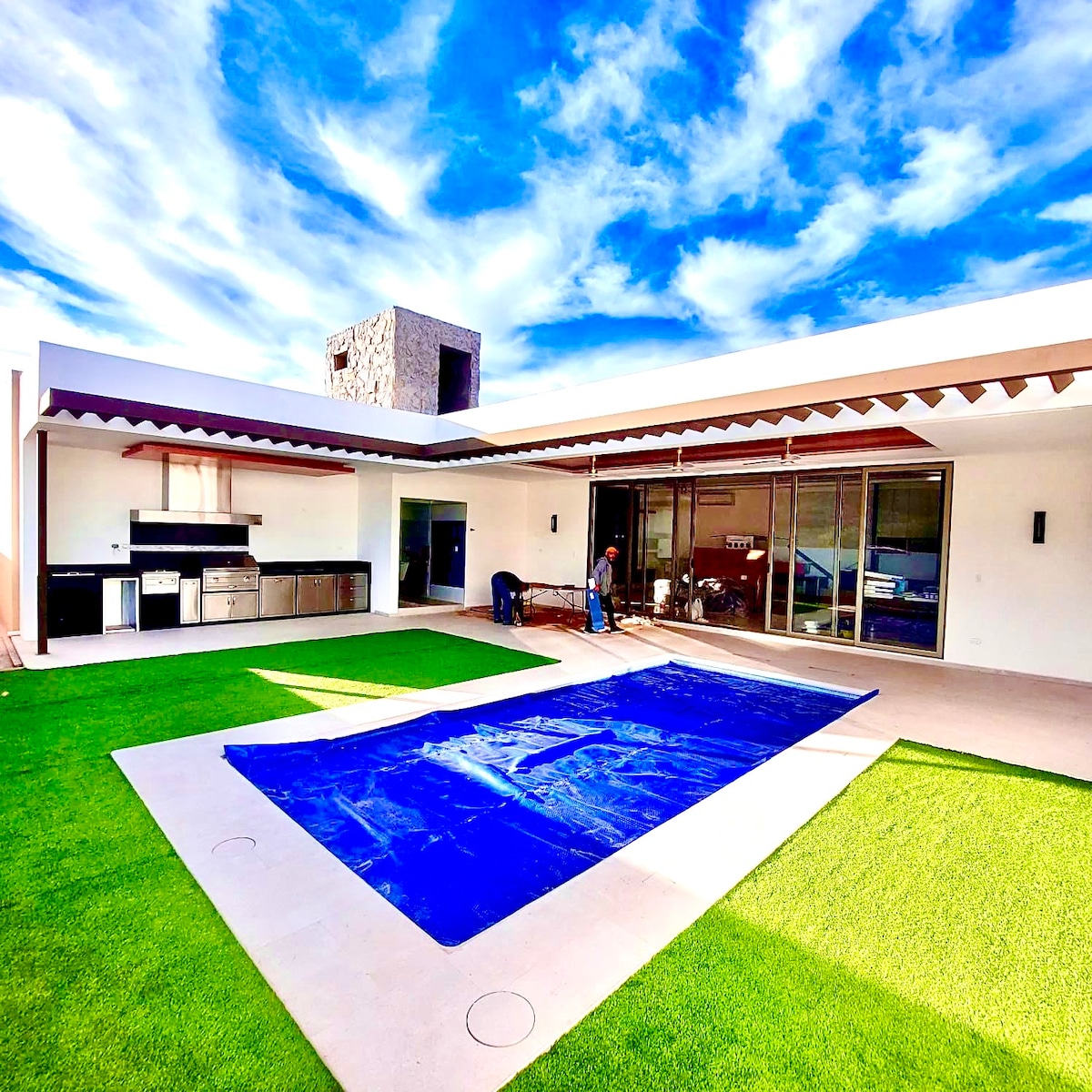 An outdoor space is showcased, featuring a smooth patio with a blue pool cover in the foreground. A well-equipped outdoor kitchen area is visible, complemented by an expansive lawn. Large glass doors reflect natural light, connecting the indoor and outdoor areas.