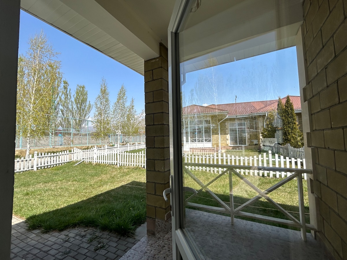 A view from the door shows a well-maintained outdoor area with a green lawn bordered by white fencing. Tall trees and the clear blue sky are visible in the background, creating a serene outdoor environment adjacent to the summer house.