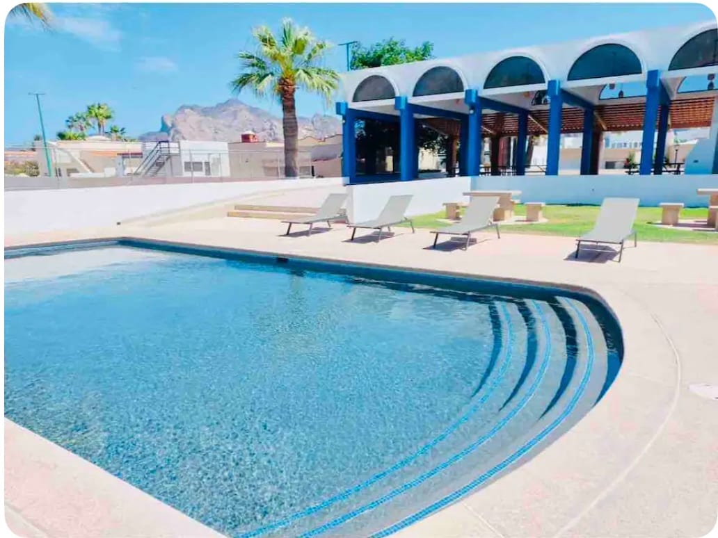 A clear swimming pool is depicted, surrounded by lounge chairs. In the background, palm trees and a shaded seating area under arched structures can be seen, with distant mountains framing the horizon.