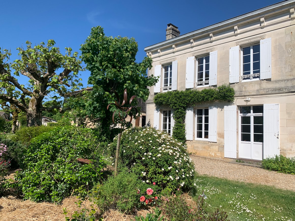 A charming stone house is framed by lush greenery and colorful flower beds. White shutters adorn the windows, while a tree provides shade nearby. A decorative feature stands in the foreground, created from natural materials, enhancing the garden's rustic appeal.