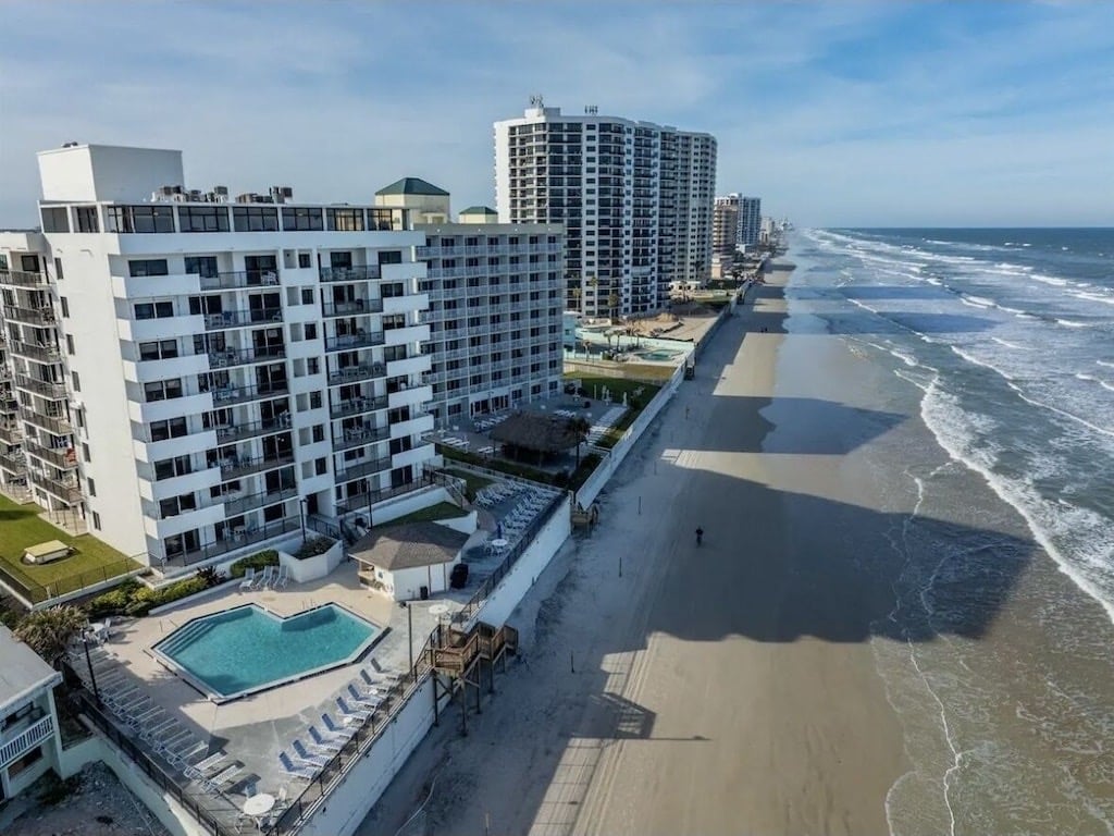 An aerial view captures the condo building alongside the beach, featuring a pool area surrounded by lounge chairs. The expansive shoreline stretches towards the horizon, with tall buildings visible in the background. The water meets the sandy beach, creating a serene coastal environment.