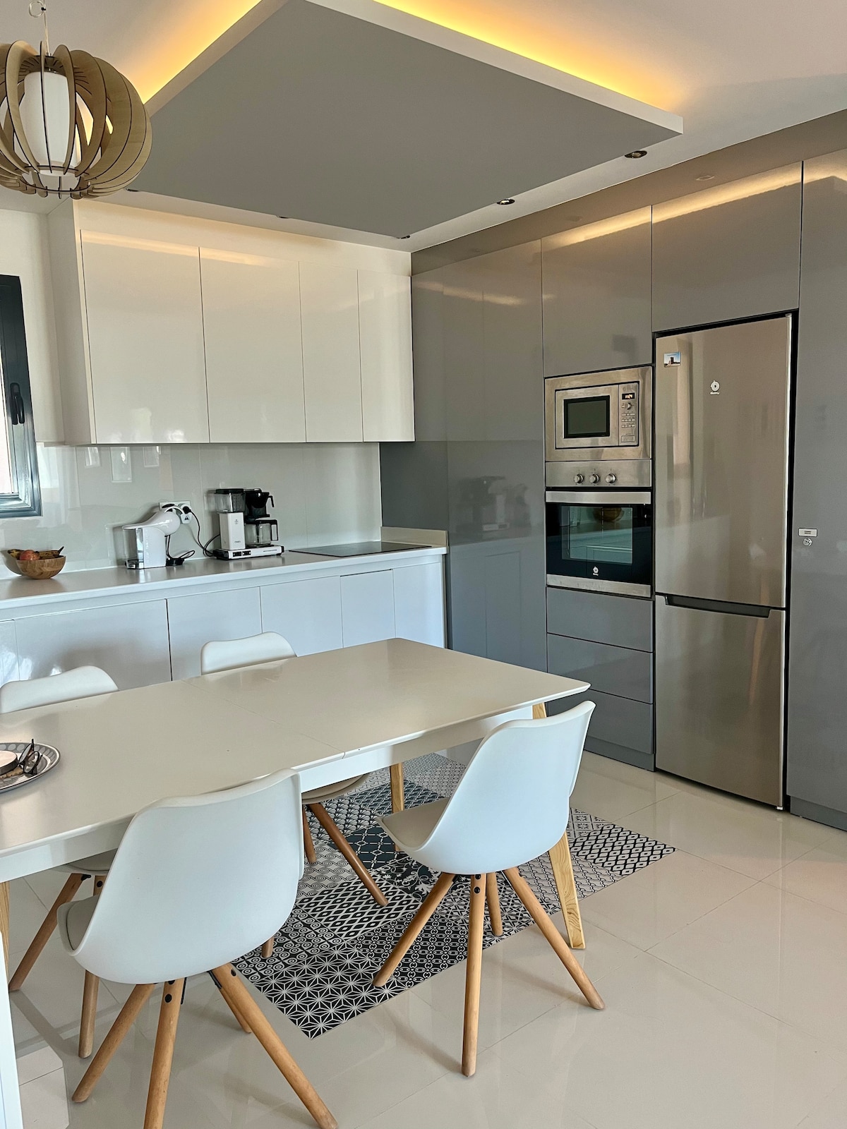 The kitchen area features a modern design with glossy cabinetry and a sleek countertop. A dining table is surrounded by four chairs, complemented by a patterned rug beneath. High-quality appliances, including an oven and coffee maker, are integrated into the cabinetry.