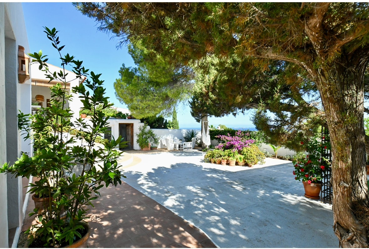 A spacious exterior area is framed by lush greenery and flowering plants. A sitting area is visible in the background, providing a tranquil space. The scene opens to views of the sky and distant coastline, enhancing the peaceful ambiance of the villa's outdoor setting.