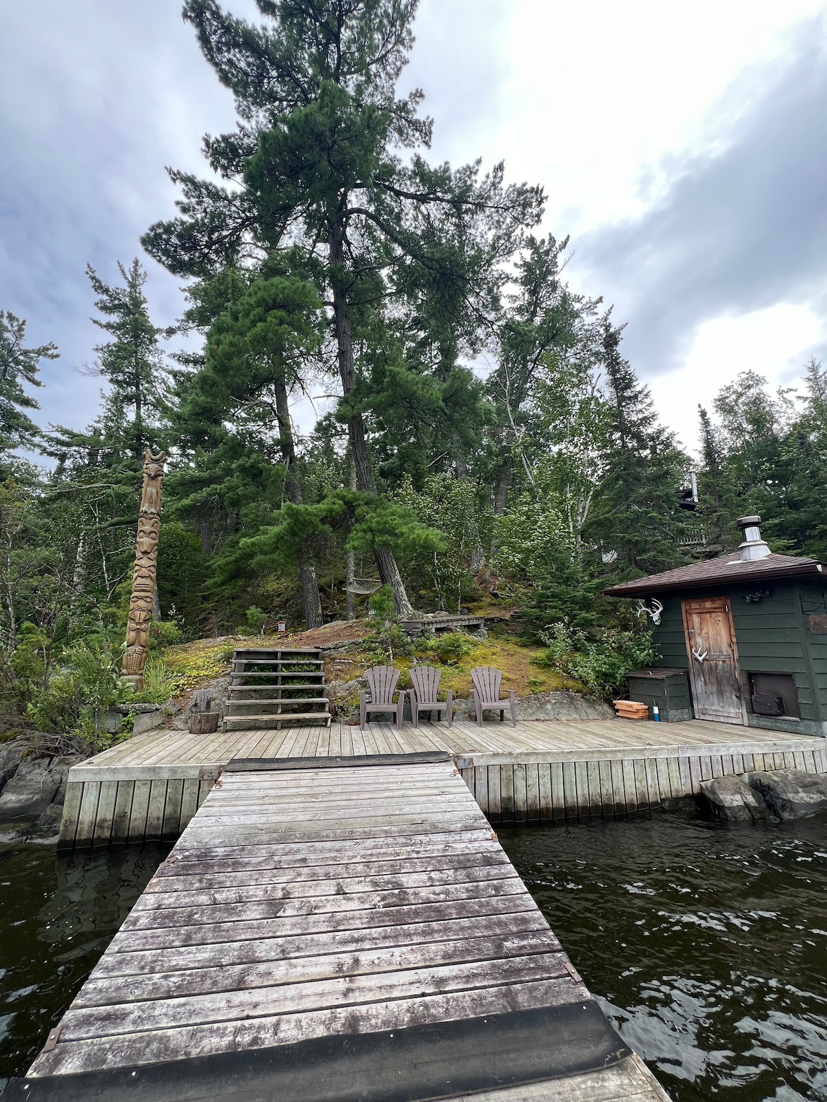 A wooden dock extends over calm water, leading to a deck area furnished with four Adirondack chairs. Surrounding trees provide a natural backdrop, while a small structure is visible to the right, enhancing the serene setting.