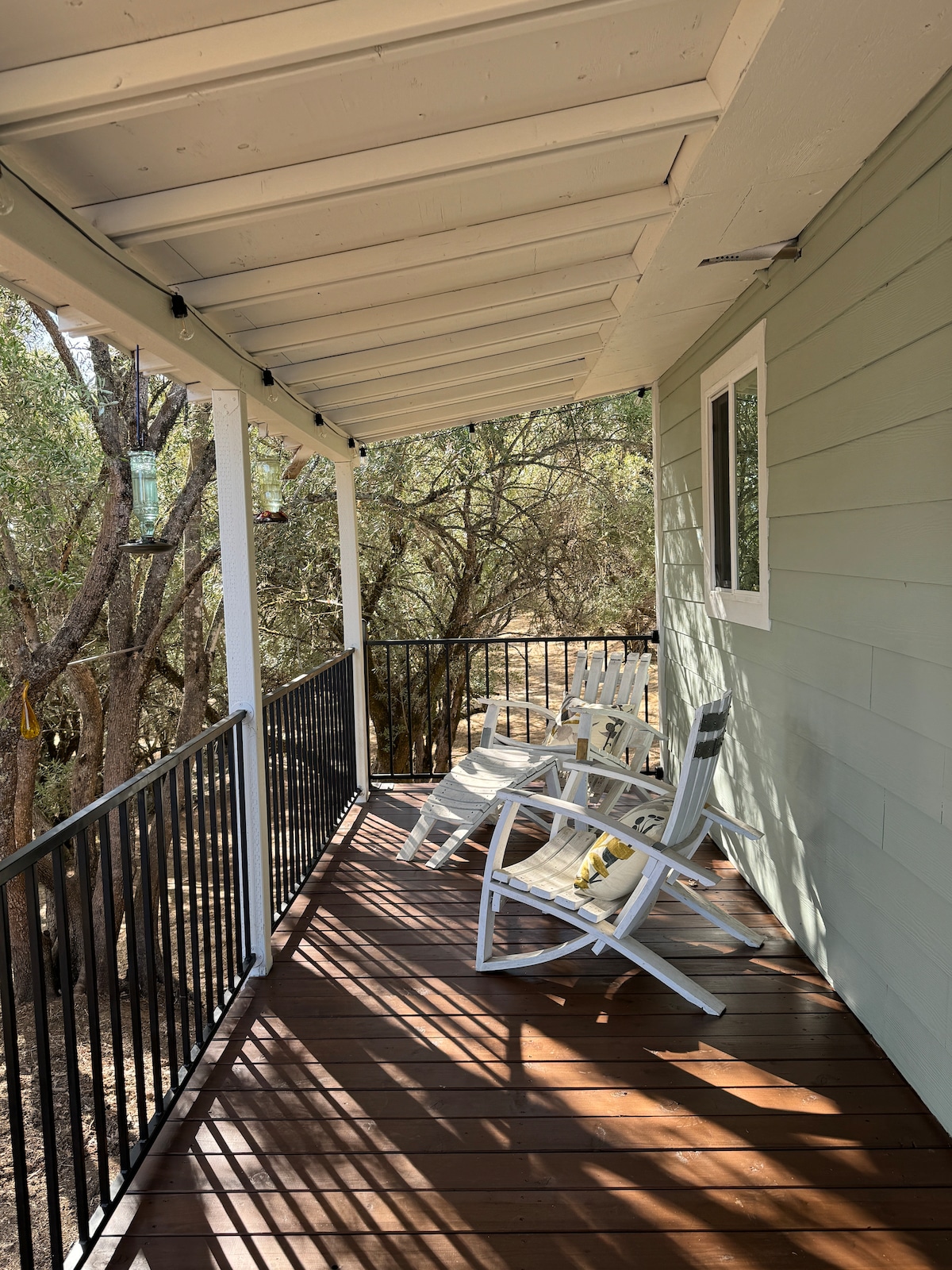 A covered porch is presented with several white rocking chairs arranged for relaxation. The wooden deck features a warm brown tone, while dappled sunlight filters through nearby trees, creating a serene outdoor atmosphere.