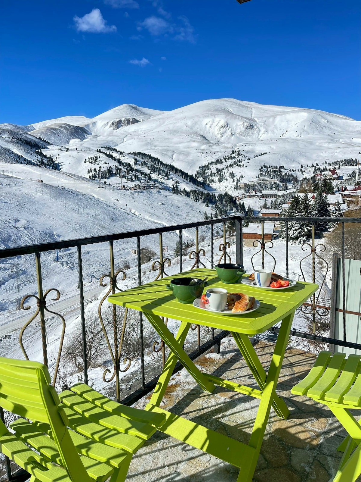 A green balcony table is set for breakfast, featuring mugs and plates, with a view of snowy mountains and clear blue skies in the background. Two matching chairs accompany the table, complementing the serene alpine landscape.
