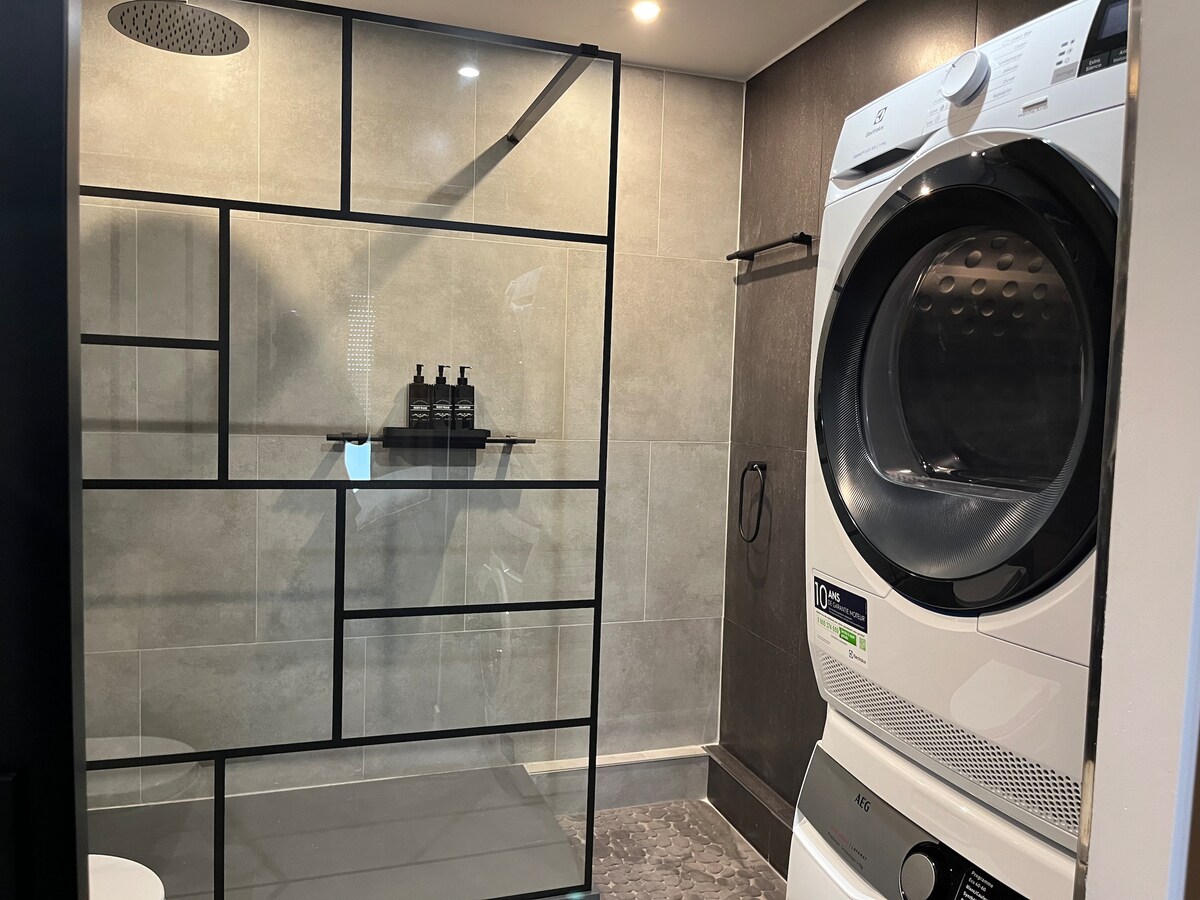 The modern bathroom features a spacious walk-in shower enclosed by a sleek glass partition. Adjacent to the shower, a washing machine is integrated into the design, and a shelf holds toiletries. Neutral grey tiles provide a contemporary backdrop.