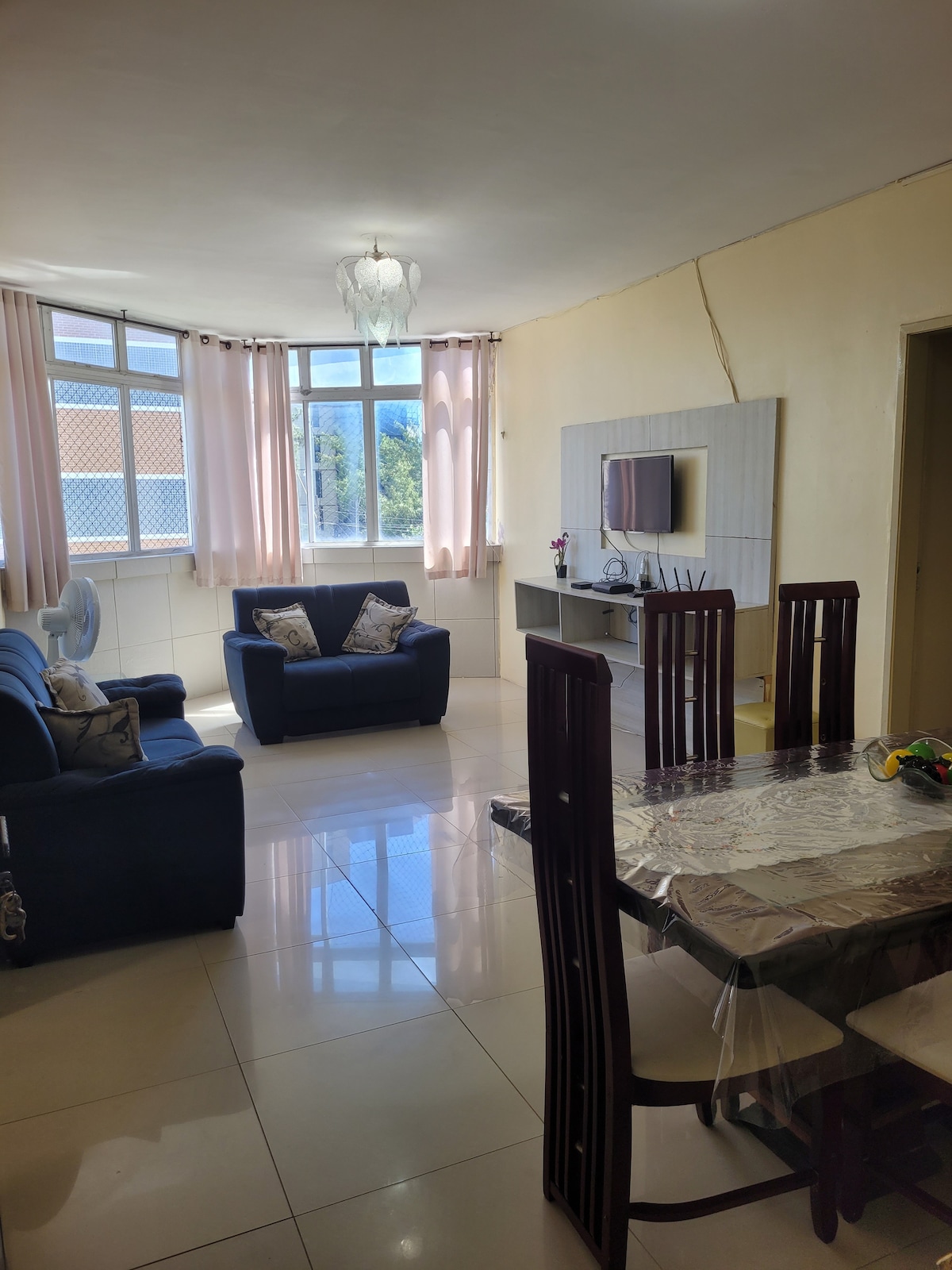 A bright living area features two sofas, one for two and another for three, arranged around a dining table set for six. Natural light enters through large windows adorned with light curtains, complemented by a TV mounted on a wall unit.