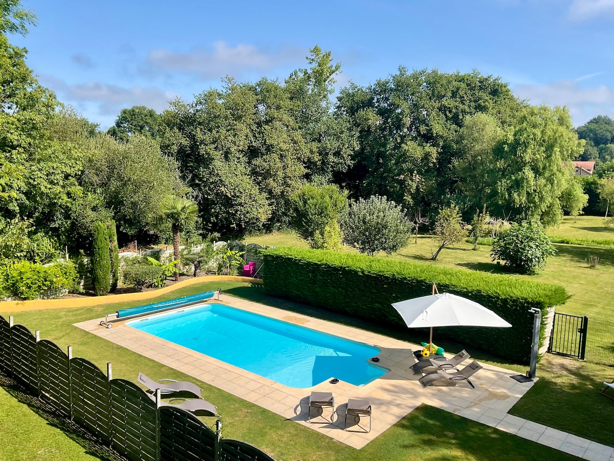 A heated swimming pool surrounded by neatly trimmed hedges is complemented by lounge chairs on the patio. An umbrella provides shade nearby, while lush greenery and trees create a peaceful backdrop, enhancing the outdoor space's tranquility.