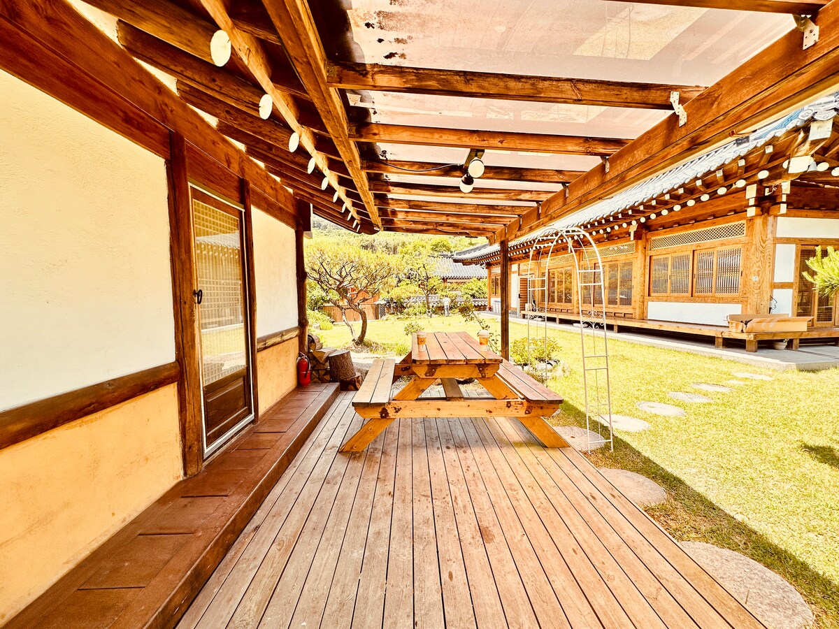 An outdoor wooden deck showcases a spacious picnic table surrounded by natural greenery. The covered area is supported by wooden beams and features string lights overhead. Nearby, traditional architecture complements the tranquil setting, inviting relaxation in a serene garden environment.