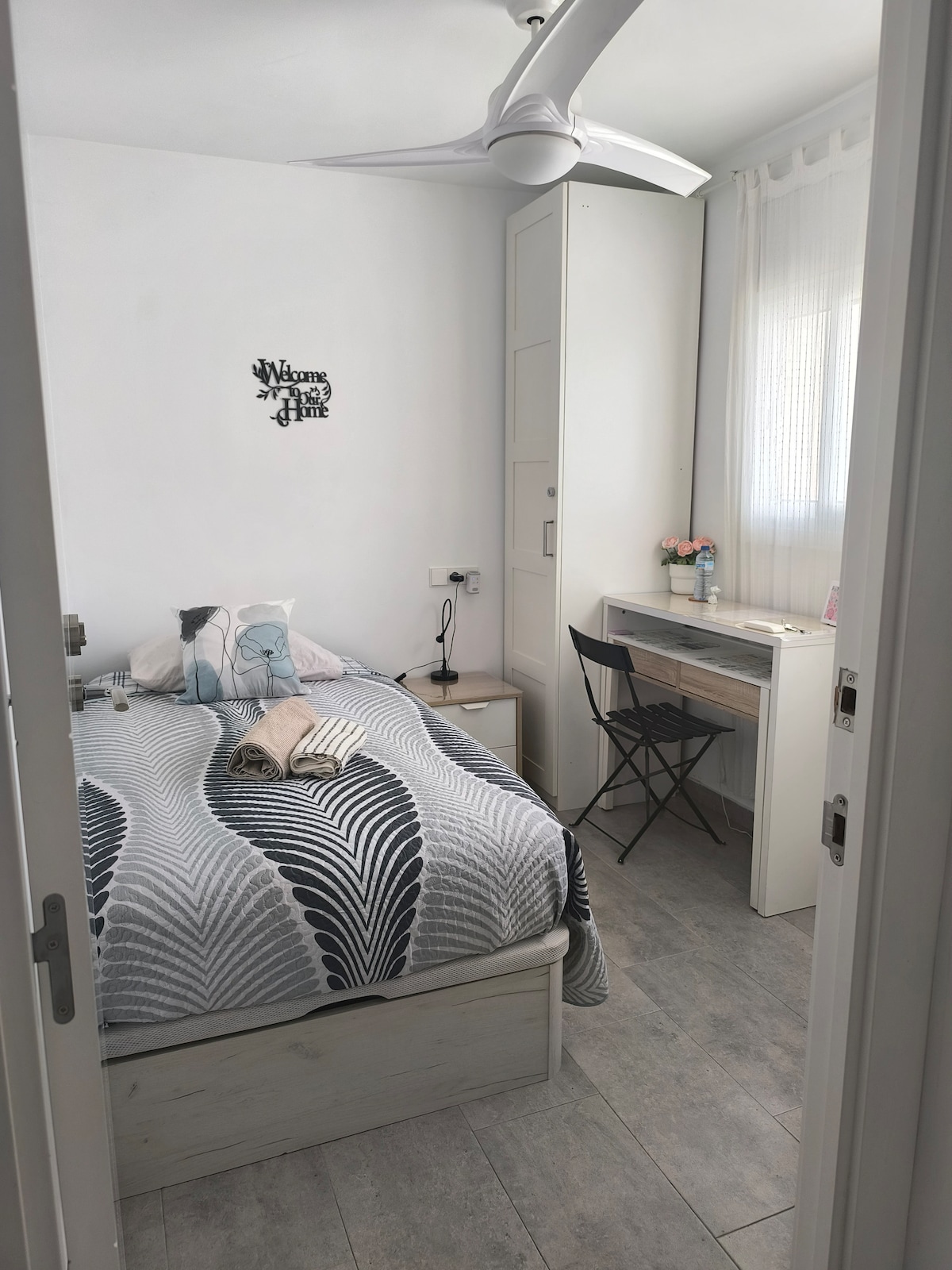 A cozy bedroom features a spacious bed with a patterned grey and white blanket. A small desk with a chair and a lamp are present, along with a decorative wall sign. Natural light filters in through a sheer curtain, creating a bright and welcoming ambiance.