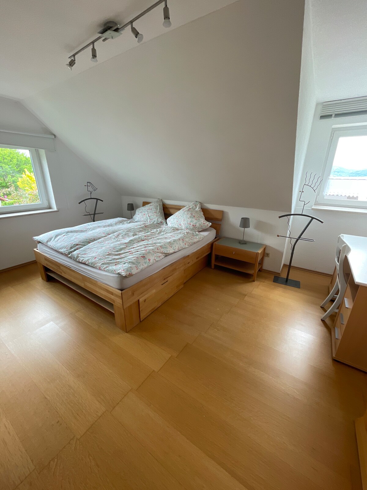 A bright bedroom features a spacious wooden bed dressed in floral bedding. Two bedside tables flank the bed, each holding a lamp. Natural light enters through two large windows, and a simple desk is tucked into the corner, enhancing the functional layout.