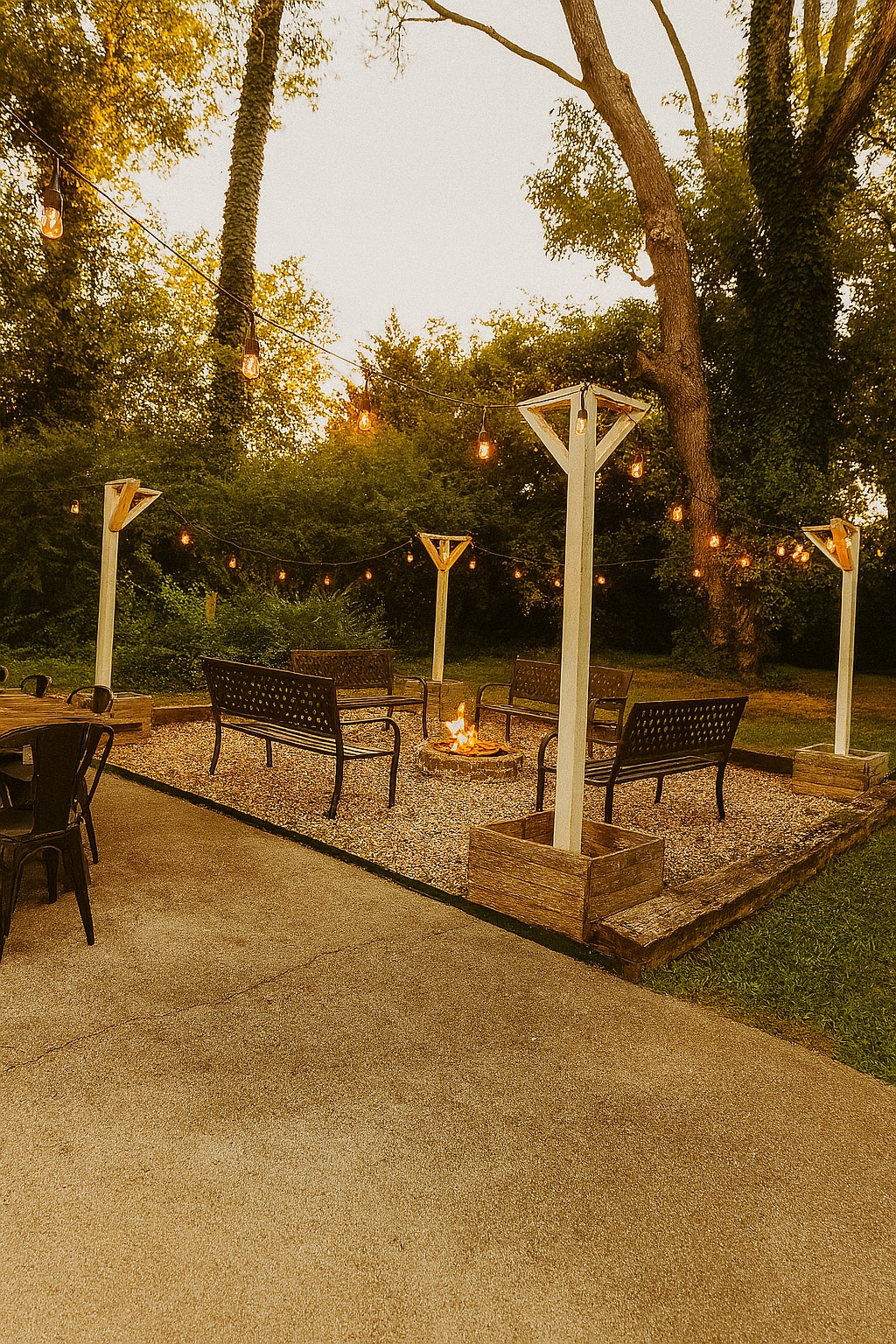 A cozy outdoor fire pit area is surrounded by comfortable seating, featuring several chairs positioned around a gravel circle. There is ambient lighting provided by Edison bulbs strung overhead, enhancing the inviting atmosphere of the backyard.