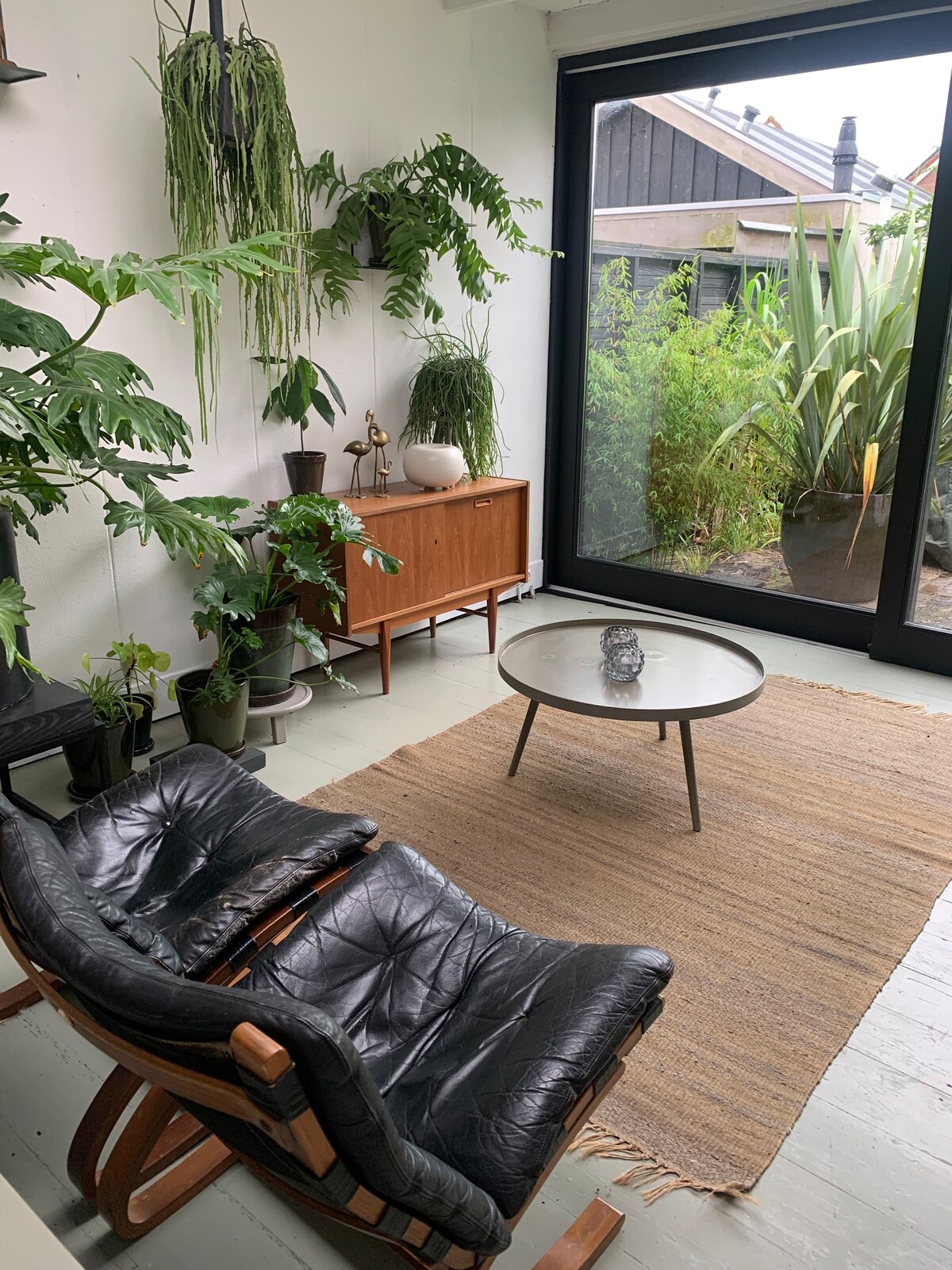 A bright corner of the room features a sleek black armchair paired with a low round table. Lush greenery surrounds the space, with various plants adorning the floor and walls. A large glass door offers a view of the garden, enhancing the connection with nature.