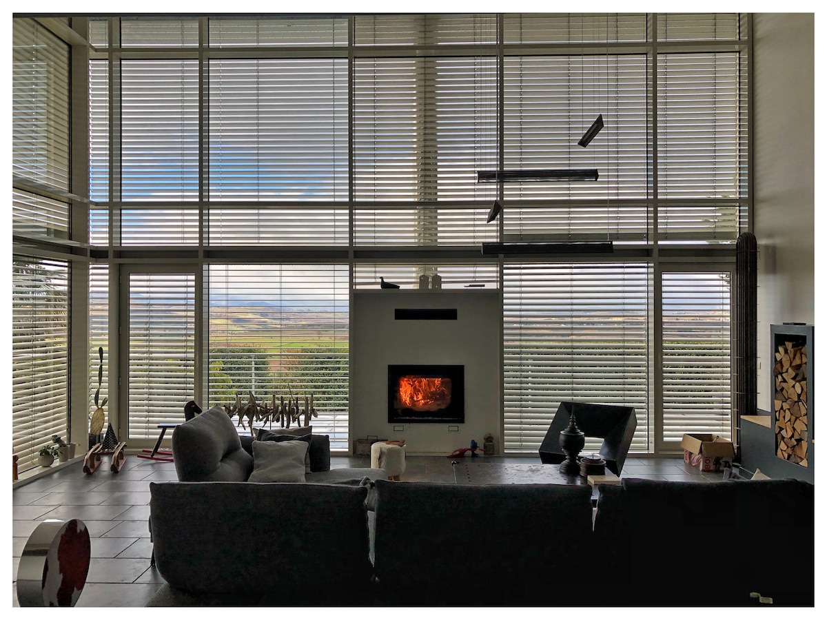 A spacious living area features large windows that offer expansive views of the surrounding landscape. A modern fireplace is centrally positioned, providing warmth. A comfortable sofa is positioned nearby, complemented by decorative items and subtle lighting fixtures hanging above.