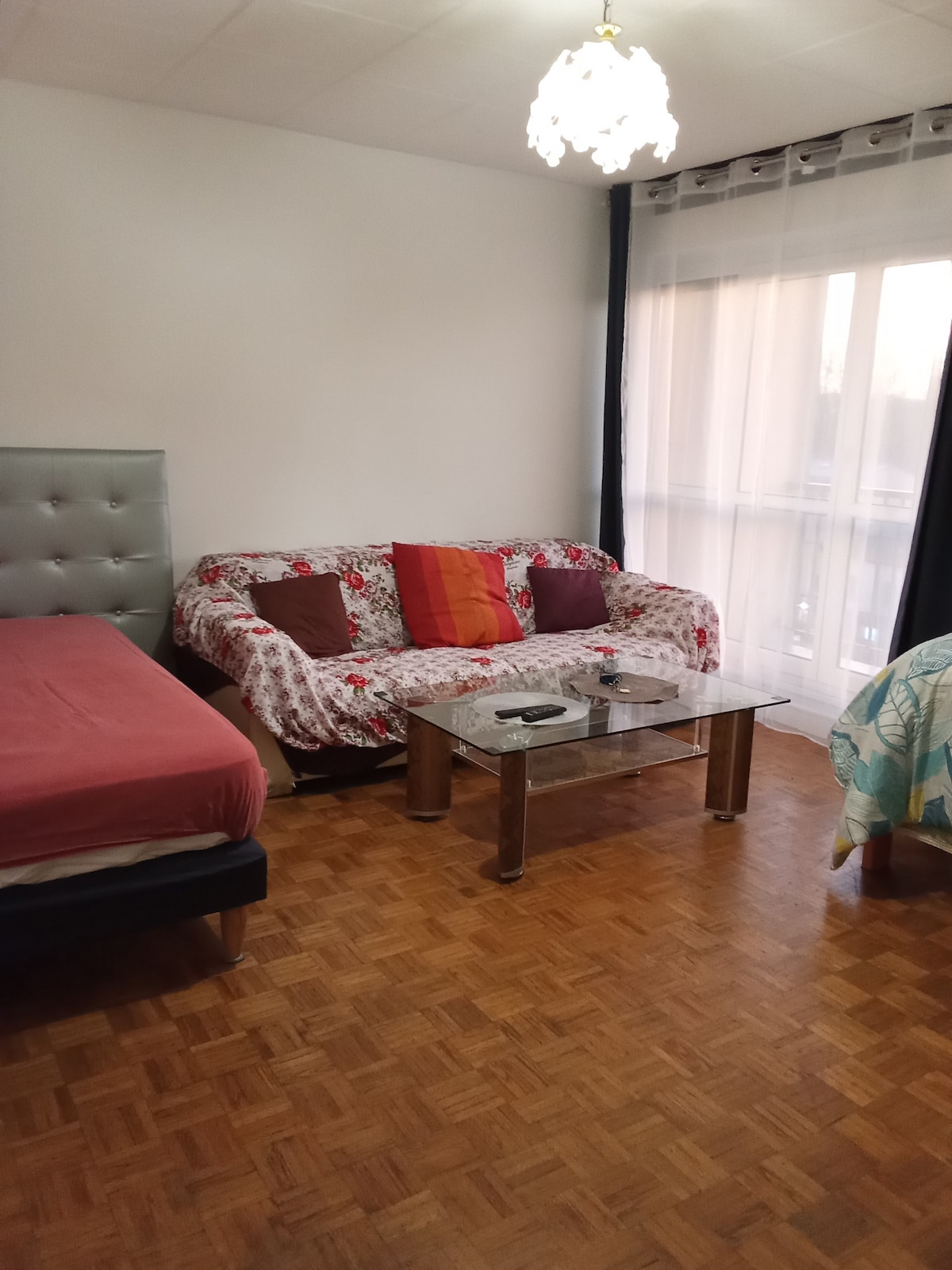 The living space features a comfortable sofa adorned with patterned cushions, paired with a glass coffee table. Two spacious beds occupy the room, and natural light flows through the large window, creating a bright atmosphere. Dark curtains frame the window, adding contrast.