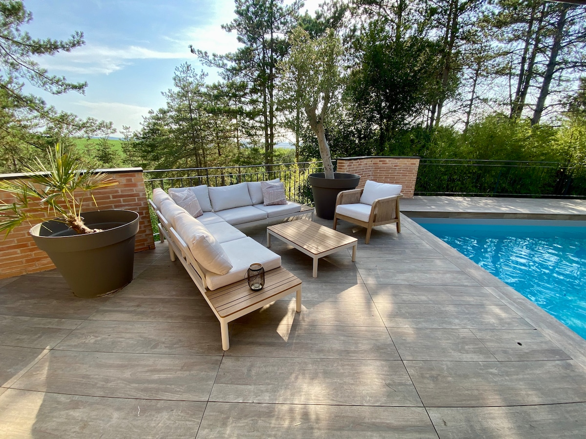 An outdoor lounge area is presented on a wooden deck, featuring a light-colored sectional sofa and a low coffee table. A potted plant adds greenery to the space. A swimming pool is visible, reflecting clear blue skies and surrounded by trees, enhancing the tranquil setting.