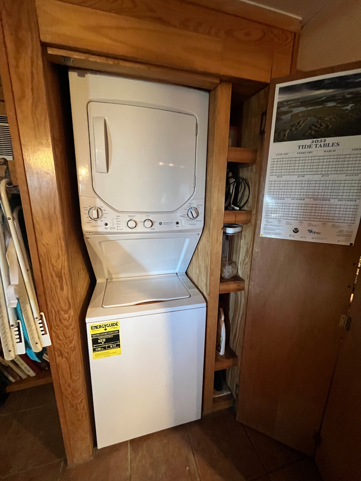 A stacked washer and dryer unit is installed within a wooden cabinet, featuring a simple, functional design. Shelving is visible alongside the unit, providing space for household items. The interior is illuminated softly, highlighting the clean lines of the appliances against the warm wood tones.