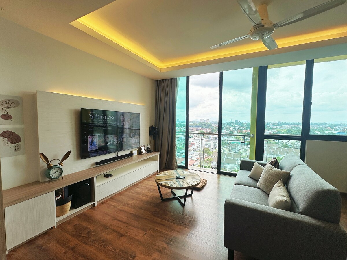 A comfortable living area features a gray sofa positioned near large floor-to-ceiling windows offering expansive views of Kuching city. A flat-screen TV is mounted on a light-colored media console, and warm ambient lighting highlights the ceiling design.