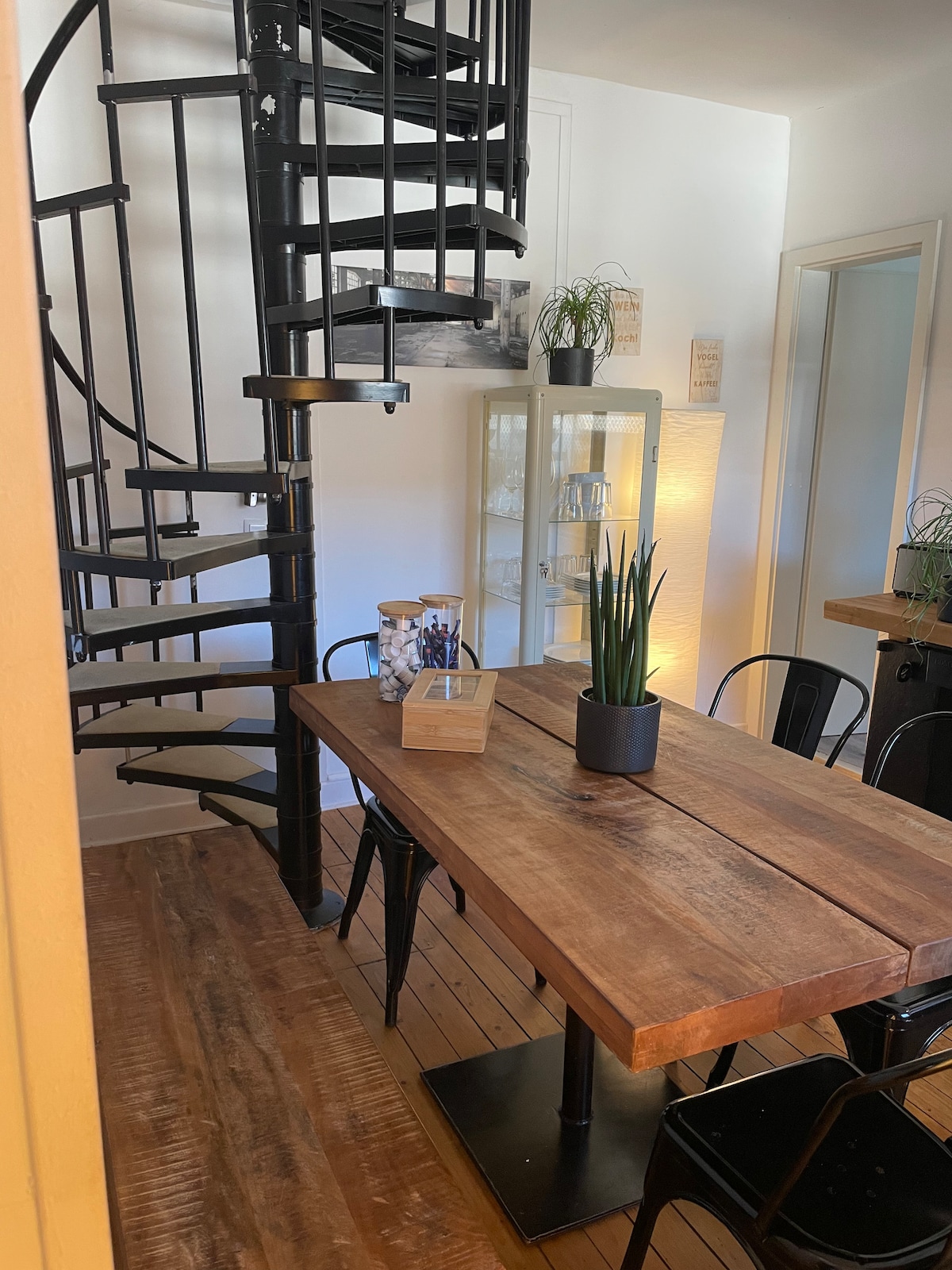 A wooden dining table with black chairs is positioned in the foreground. A spiral staircase ascends to the upper level, adding a unique architectural feature. A small potted plant and utensils are placed on the table, with soft lighting enhancing the inviting atmosphere.