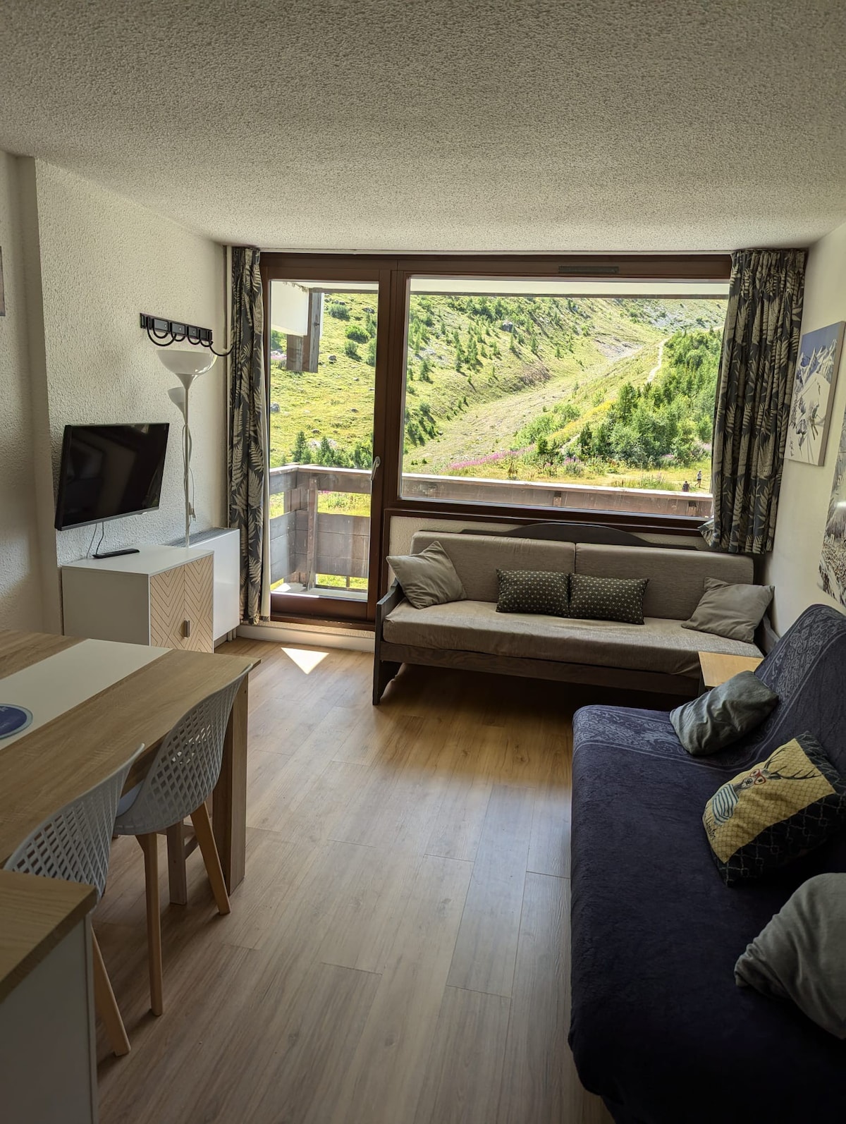 A bright living area features a large window offering views of the surrounding mountains and greenery. A spacious sofa is accompanied by a dining table with chairs. A TV is positioned against a wall, providing a functional focal point for relaxation.