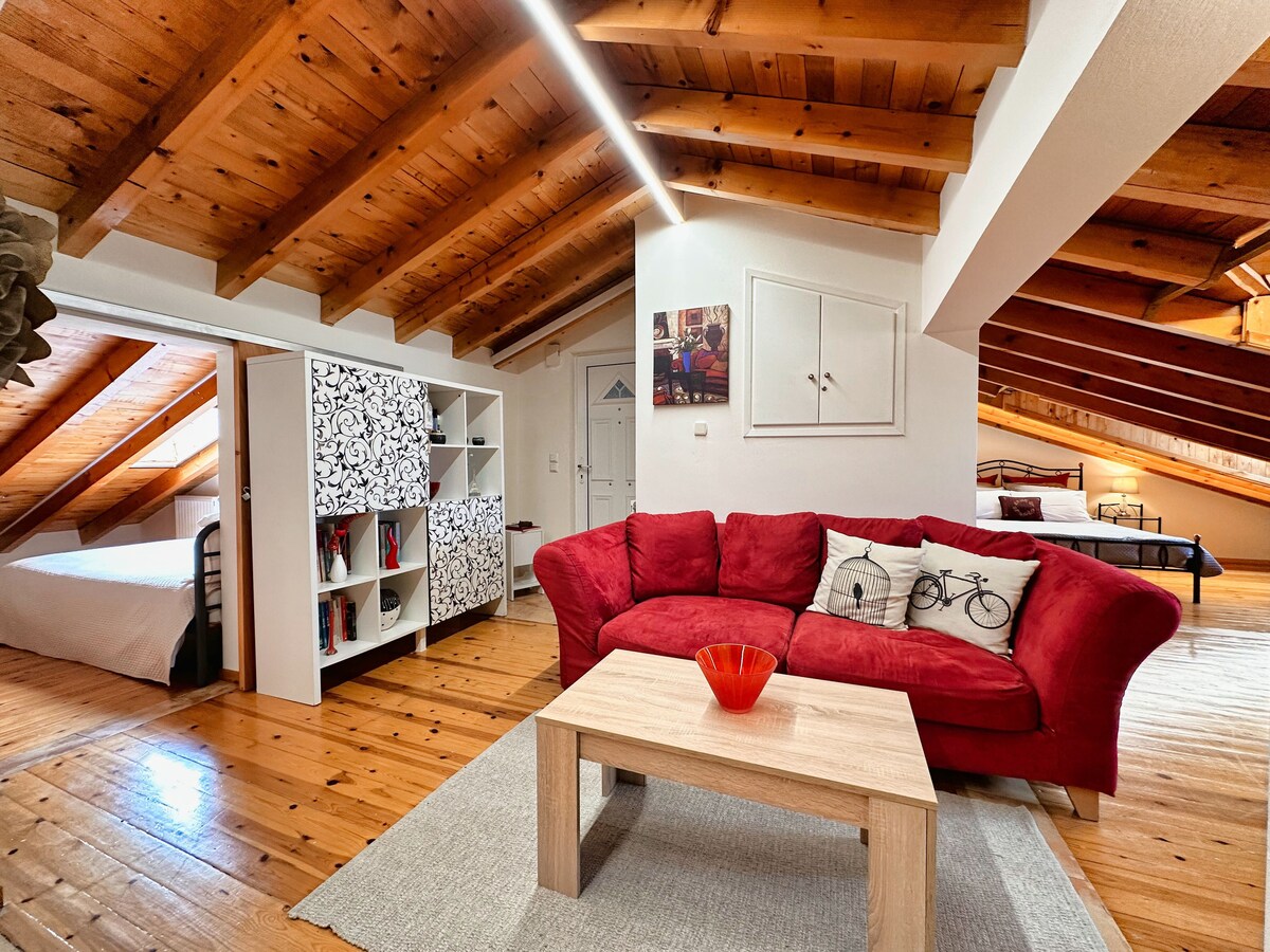 A bright living area is highlighted by wooden beams and warm tones. A red sofa with decorative cushions faces a light-colored coffee table. A bookshelf is positioned nearby, and access to a spacious sleeping area is visible in the background.