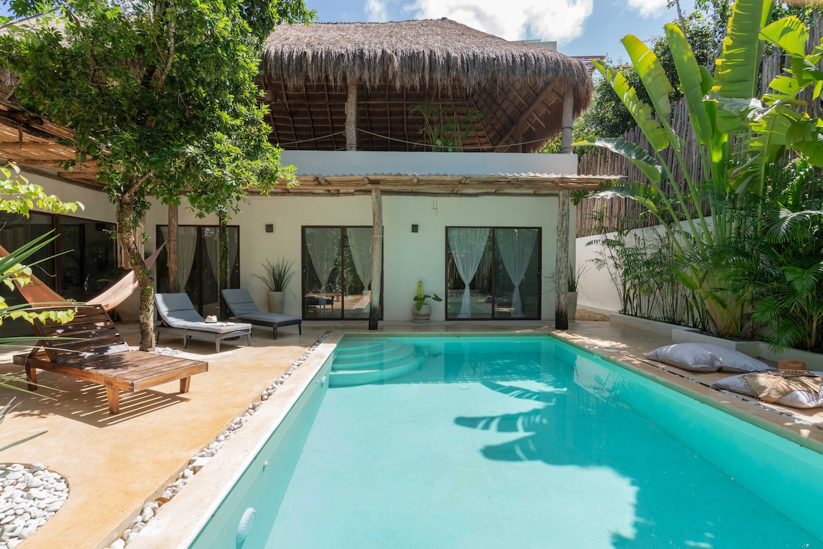 A spacious outdoor area features a private swimming pool surrounded by a sun-drenched terrace. Comfortable lounge chairs are positioned near the water, while a thatched-roof structure provides shade. Lush tropical plants border the space, contributing to the tranquil ambiance.