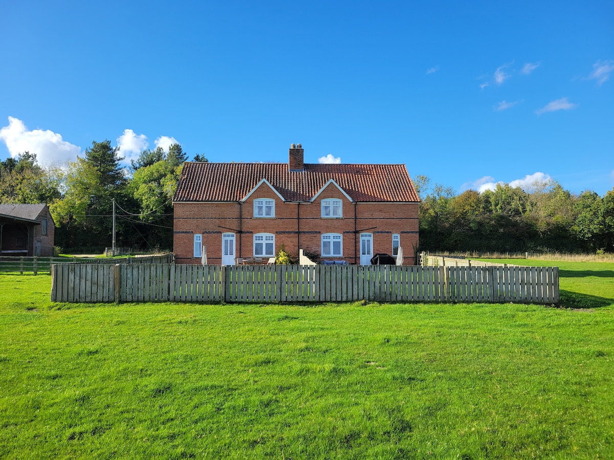 The cottage features a traditional brick exterior with a gabled roof and white-framed windows. A fenced garden surrounds the property, providing a sense of privacy. The expansive green lawn offers ample space for outdoor activities, with trees lining the backdrop for a natural setting.