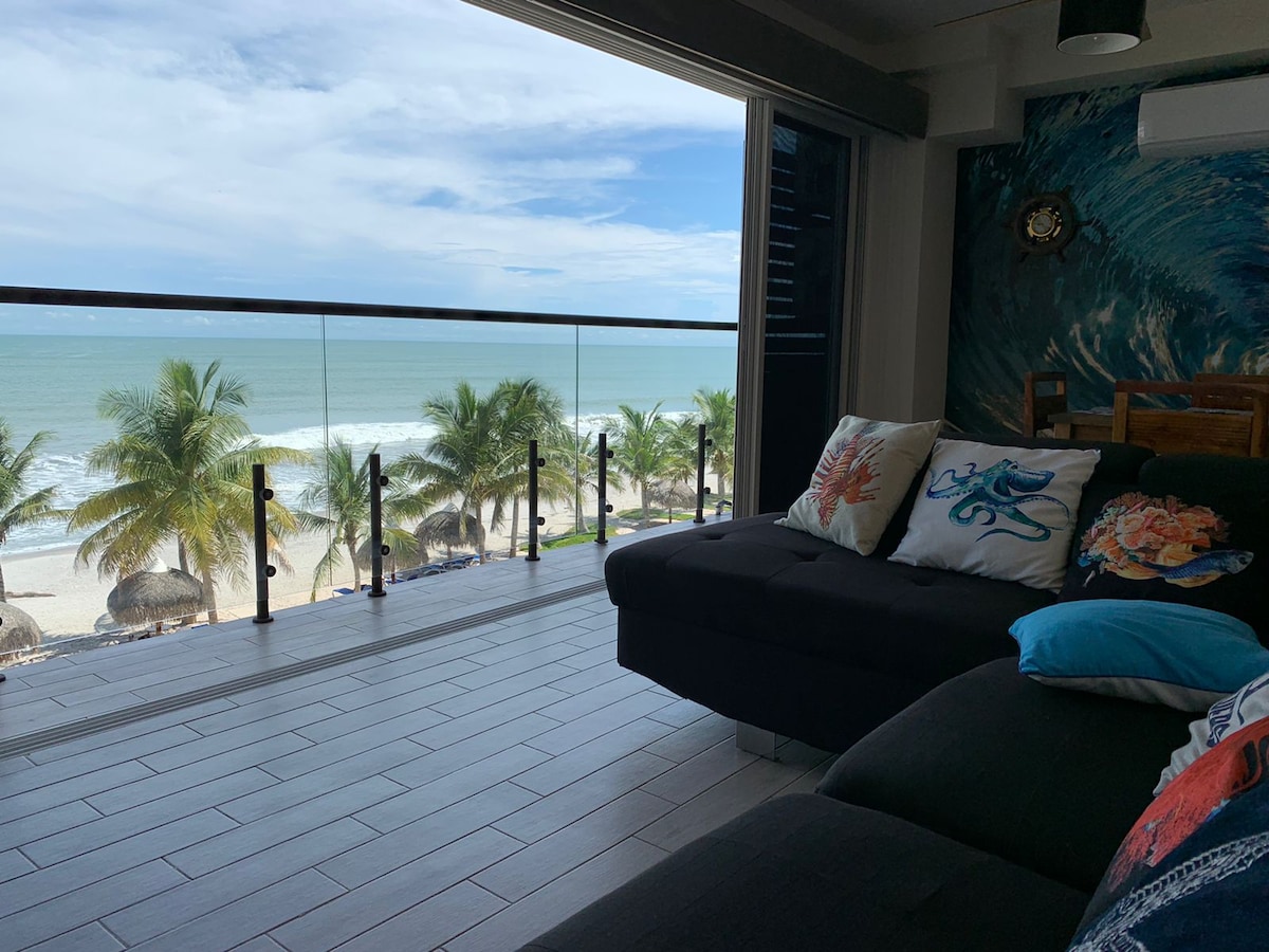 A spacious living area features a dark sectional sofa adorned with decorative pillows. Large glass doors provide direct views of the beach, with palm trees visible along the shoreline. Light colors dominate the walls, adding to the bright ambiance created by natural light.