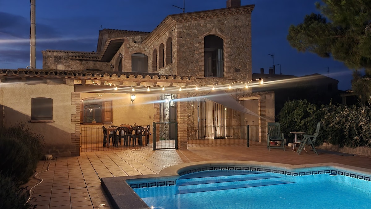 A stone house with ample outdoor space is illuminated by soft lights in the evening. The pool glimmers invitingly, while the patio features a large dining table surrounded by chairs. A boundary fence separates the pool area from the garden, adding privacy.