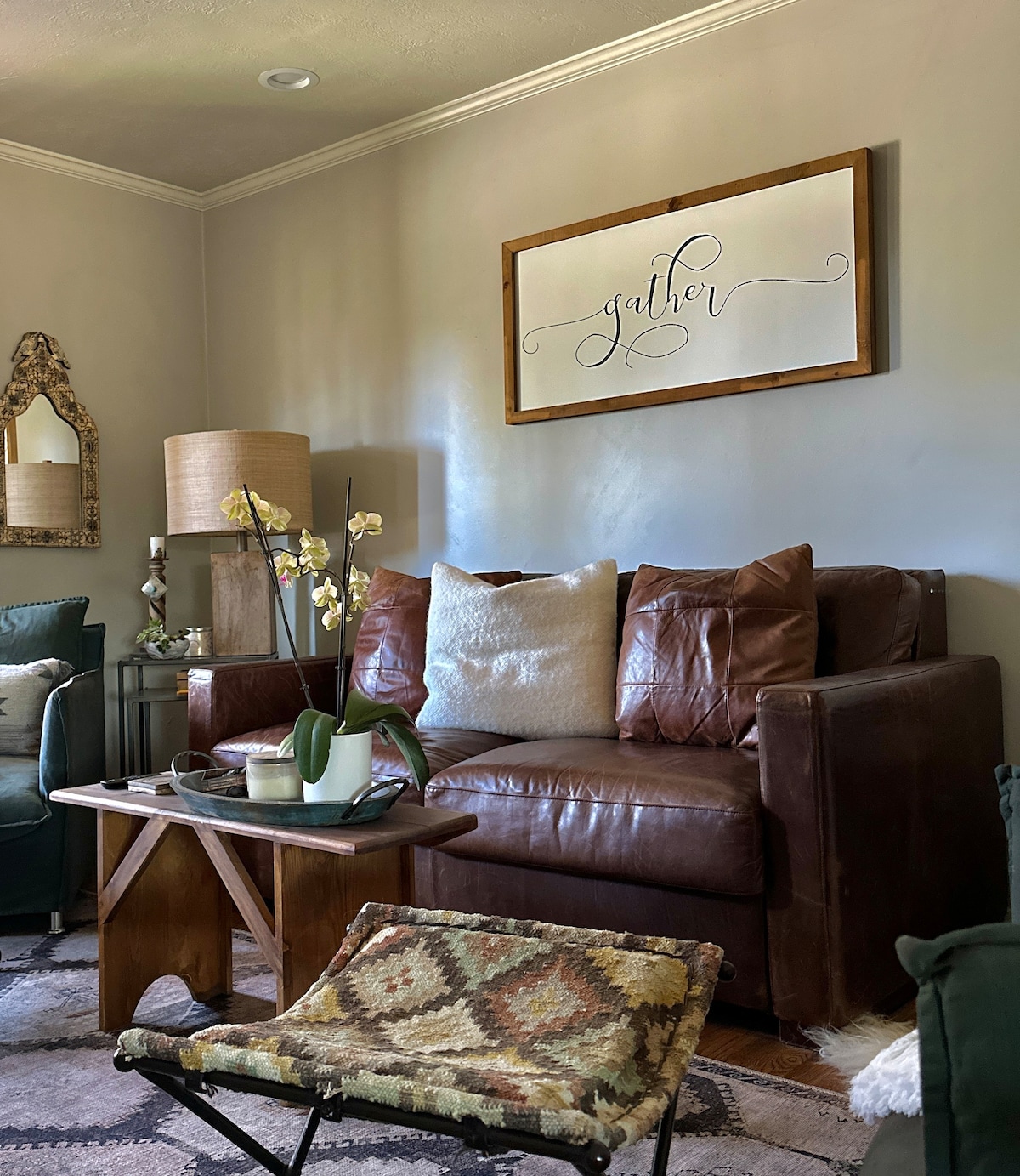 A cozy seating area features a brown leather sofa adorned with cushions and a decorative wall hanging that reads 'gather'. A small wooden side table holds a potted plant and a book, while a patterned ottoman adds additional seating.