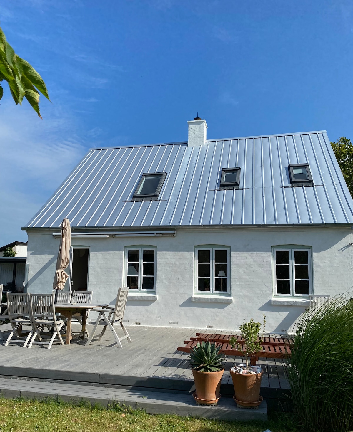 The exterior of a two-story white house is presented, featuring a metal roof and multiple windows. A spacious wooden deck is visible, furnished with a dining table and chairs. Green plants and a parasol are positioned nearby, enhancing the outdoor dining area.