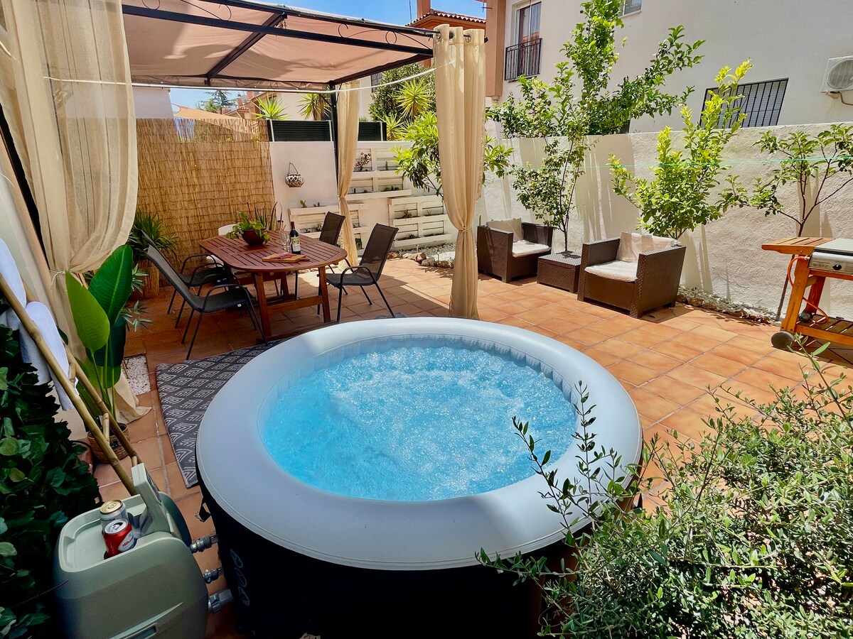 A private patio features an inflatable hot tub surrounded by greenery and sunlit tiles. Comfortable seating areas are arranged, including a dining table with chairs and shaded lounges. The space is designed for relaxation, enriched by natural light filtering through soft curtains.