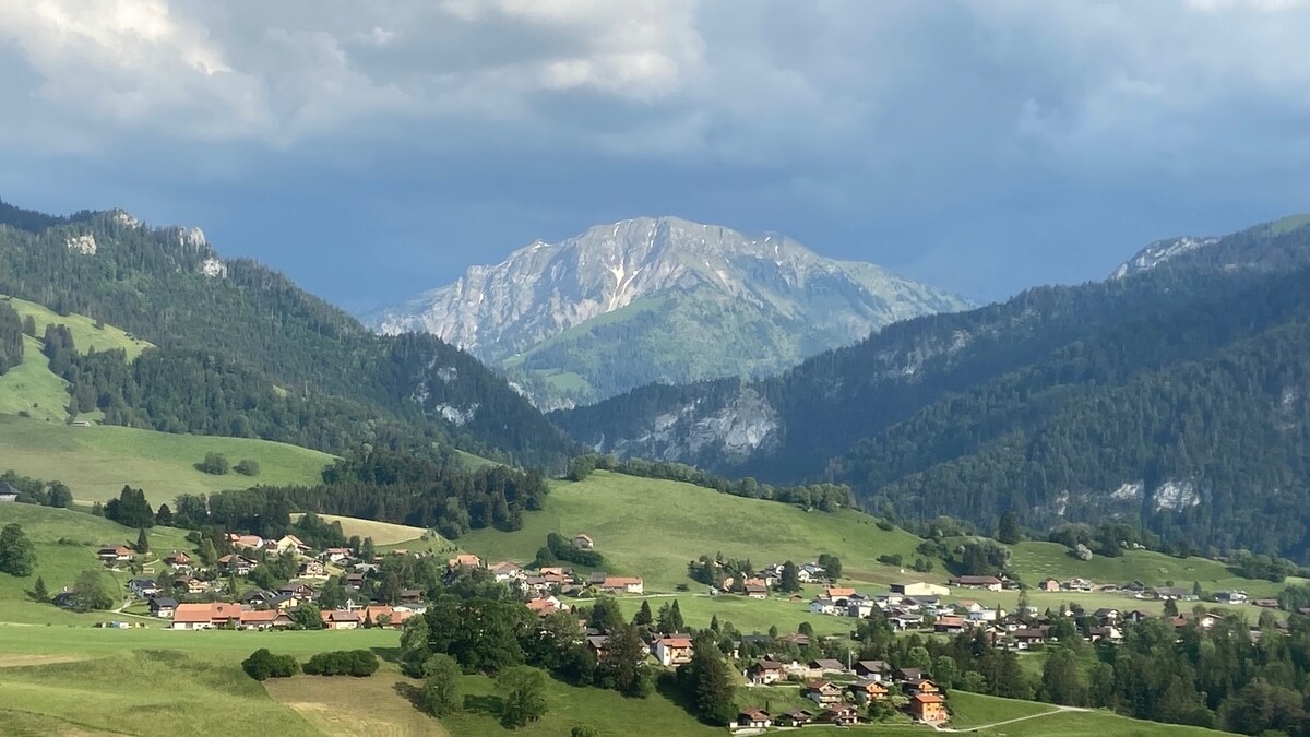A broad view captures the surrounding countryside, showcasing rolling green hills and a distant mountain range under a cloudy sky. The village below is partially visible, with several houses scattered across the landscape, emphasizing the serene rural environment.
