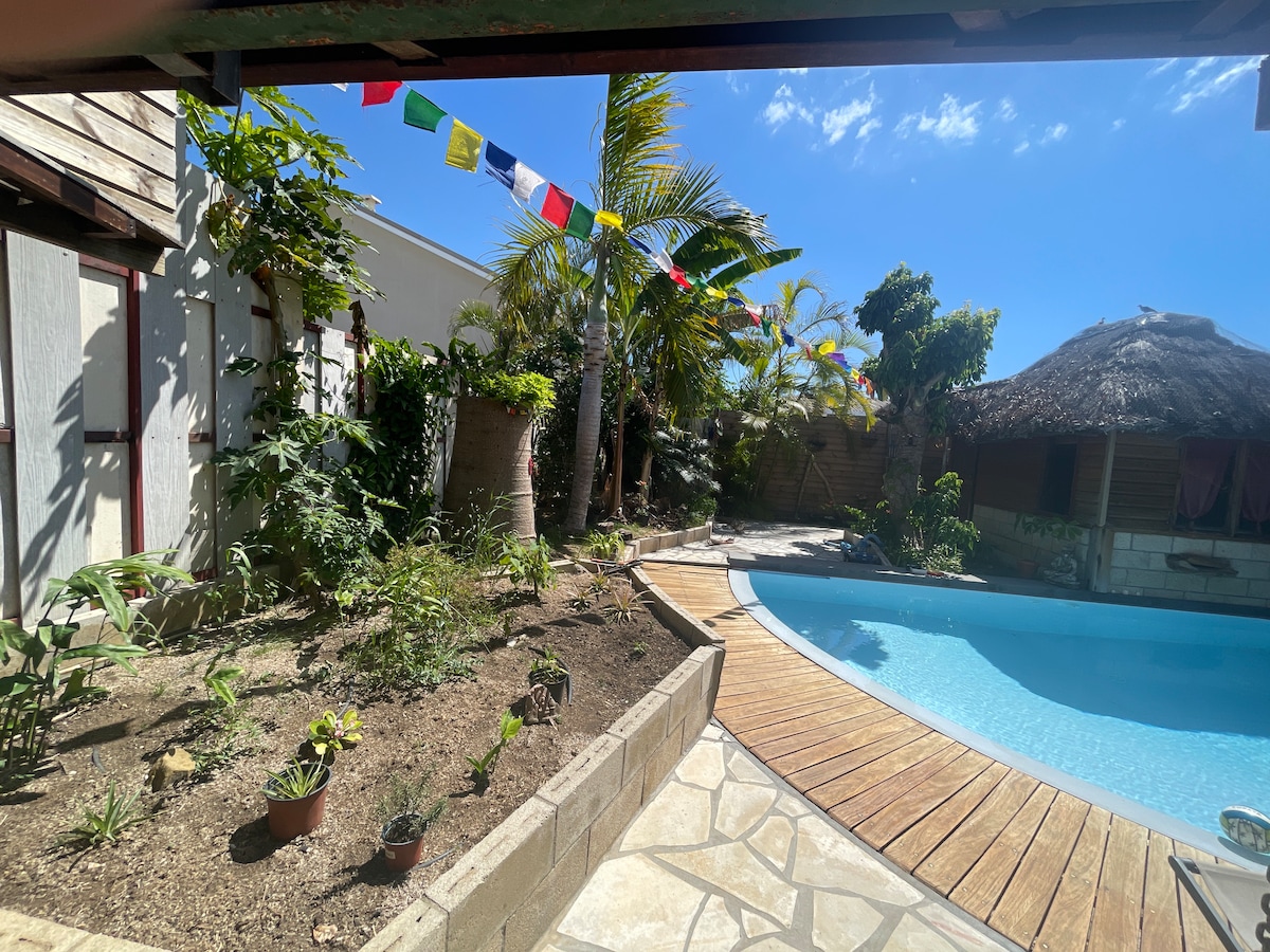 A serene outdoor area features a turquoise pool surrounded by wooden decking and tropical plants. Colorful flags are strung overhead, gently fluttering in the breeze. A thatched-roof structure adds to the cozy atmosphere, complementing the lush greenery in the garden.