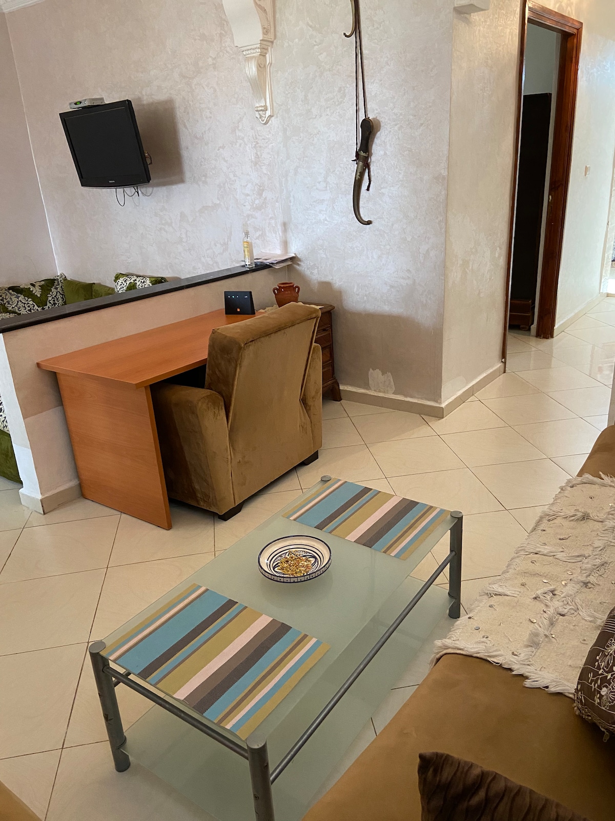 A living area is presented, featuring a small glass coffee table with decorative items. A comfortable sofa is visible, alongside a chic chair and a wooden desk. A wall-mounted television can be seen, providing a cozy atmosphere for relaxation.
