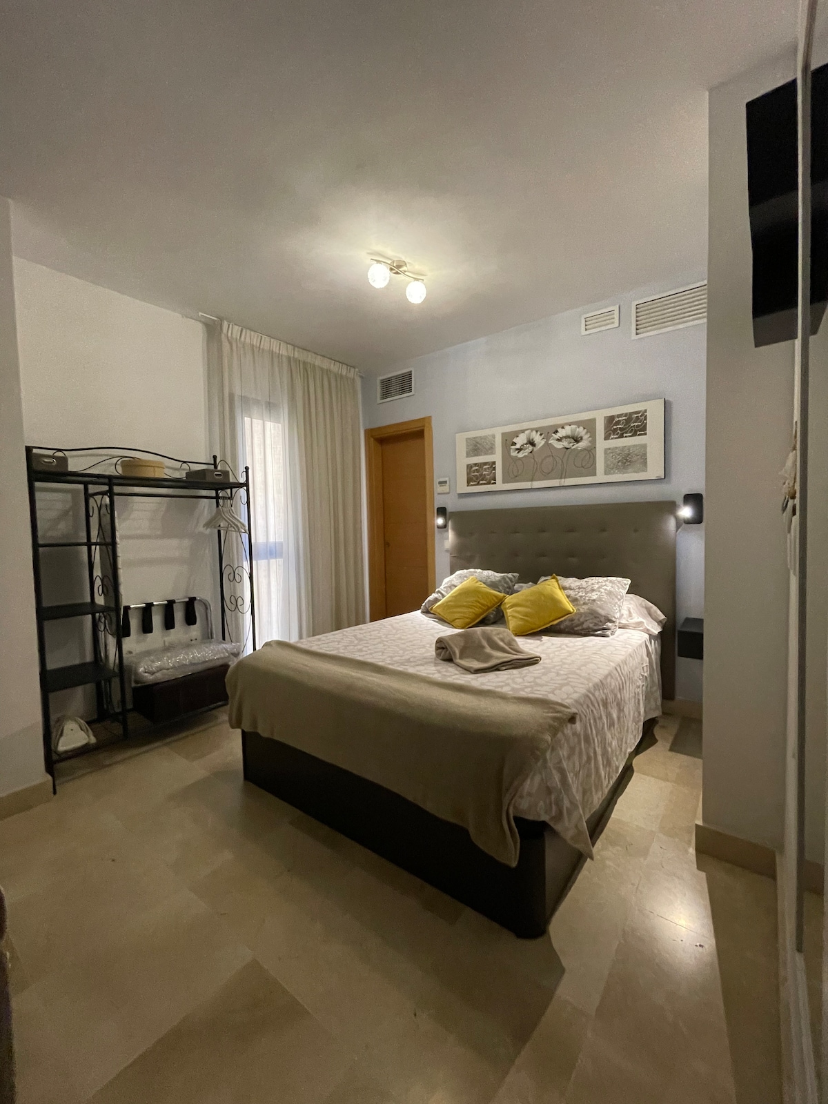 A cozy sleeping area is equipped with a spacious bed, featuring a light-colored quilt and decorative yellow pillows. A wall-mounted mirror reflects the room's layout, while natural light filters through sheer curtains. A clothing rack and a doorway are visible, adding to the room's functionality.